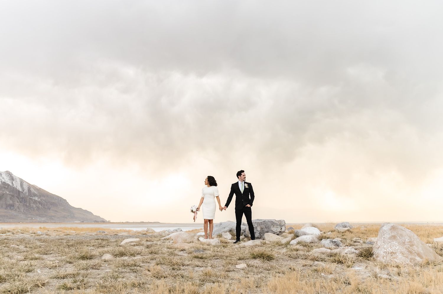 Couple holding hands on rocky terrain with dramatic cloudy sky and mountain backdrop during outdoor engagement session.