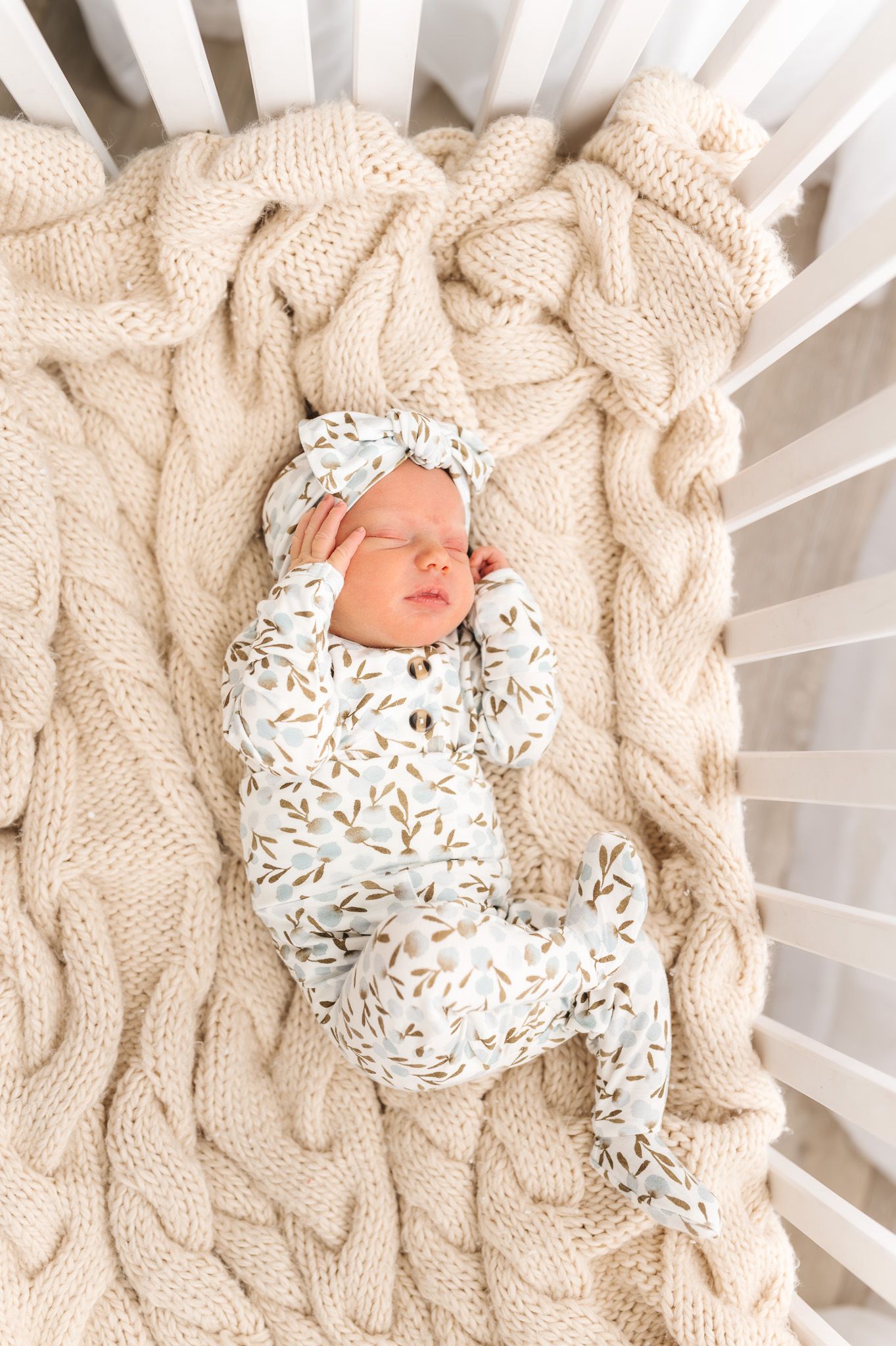 Sleeping newborn baby in white and gold floral outfit and matching bow headband on cream cable knit blanket in crib.