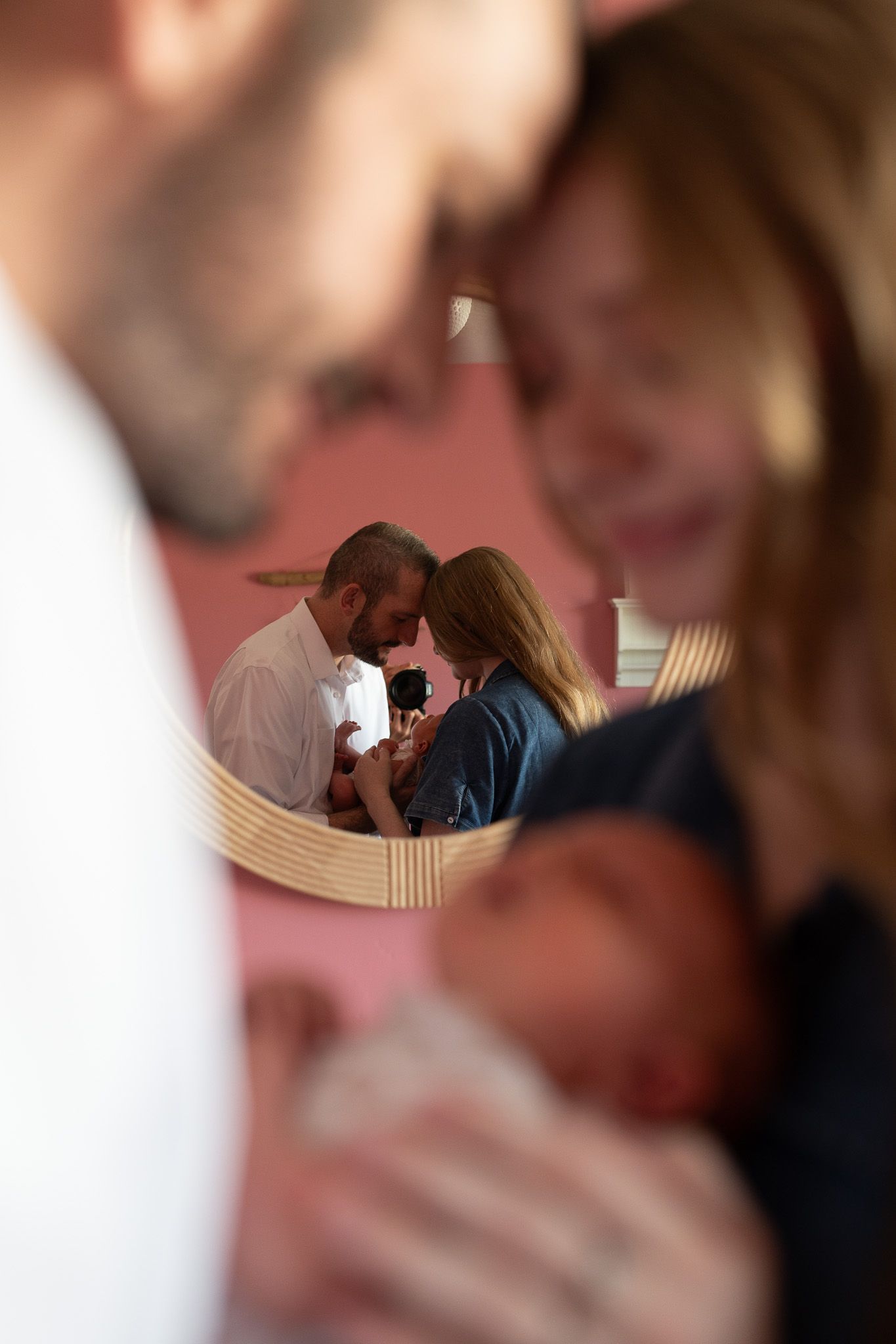 Couple touching foreheads while holding newborn baby, reflected in round mirror with pink wall background.