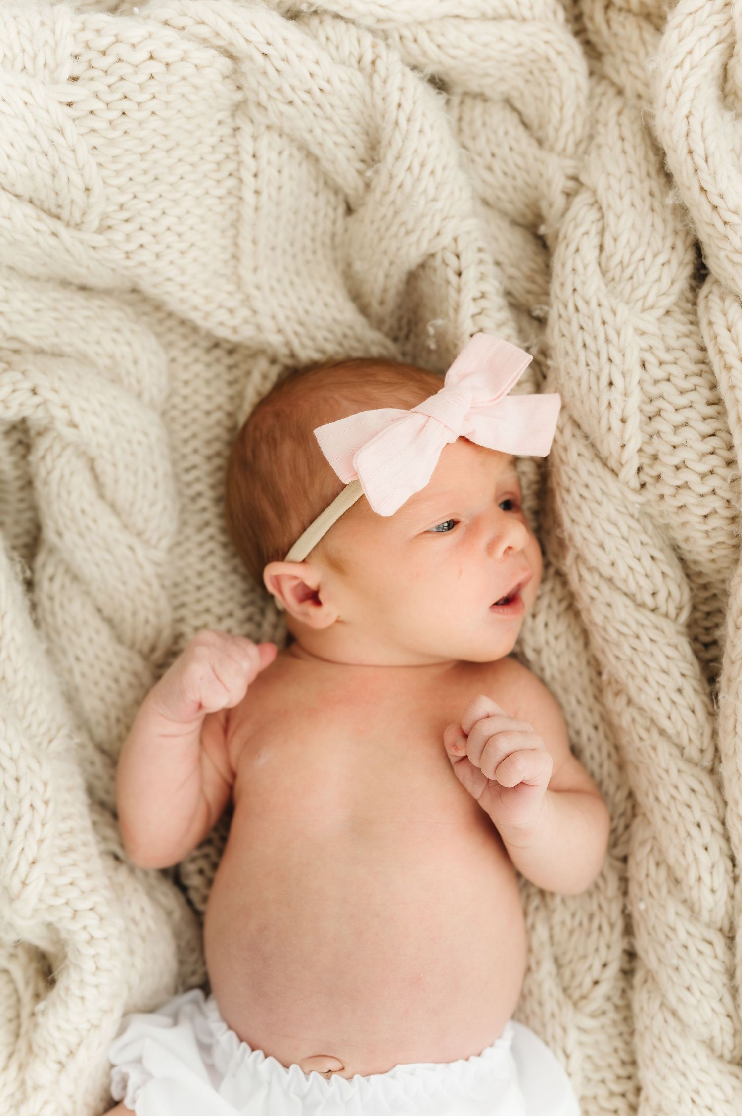 Newborn baby wearing pink bow lies on cream cable knit blanket during portrait session.