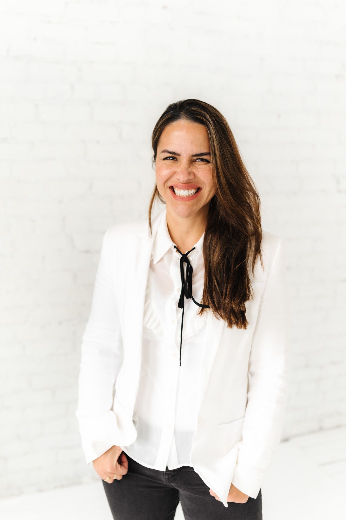 Professional in white blazer and black tie smiles against minimalist white background.