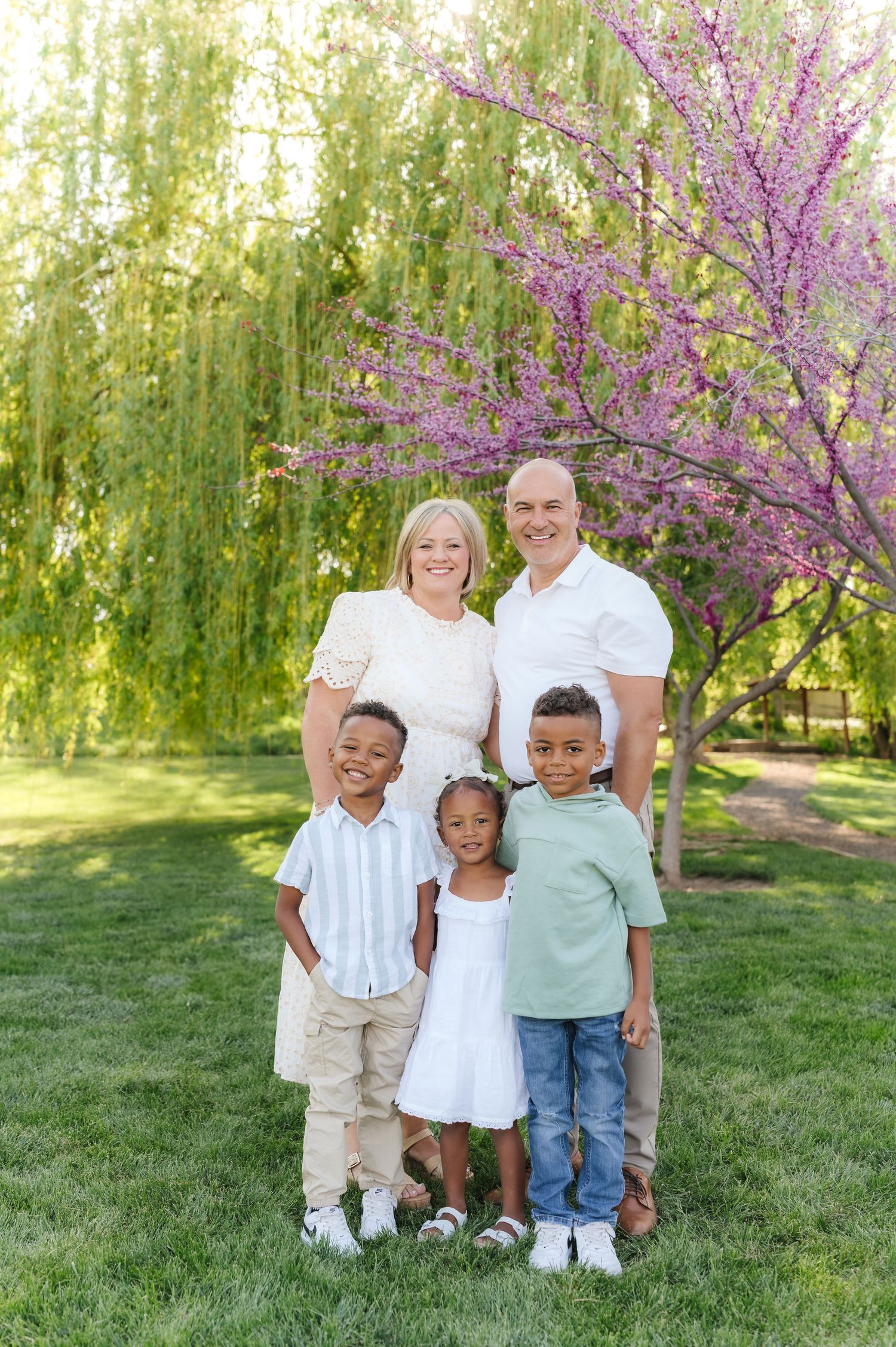Family portrait in white outfits standing under blooming cherry tree in spring garden setting.