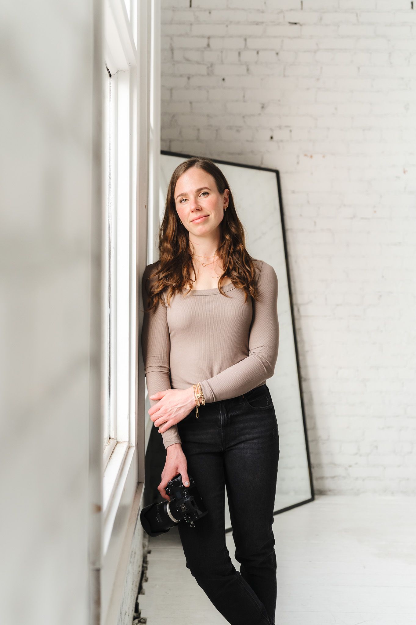 Woman photographer holding camera leans against white brick wall near window in bright studio space.