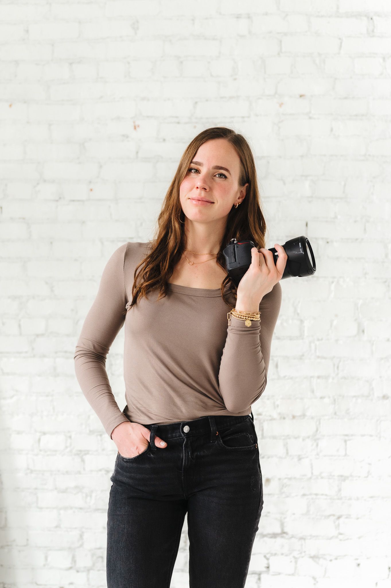 Professional photographer in neutral top holding camera against white brick wall background.
