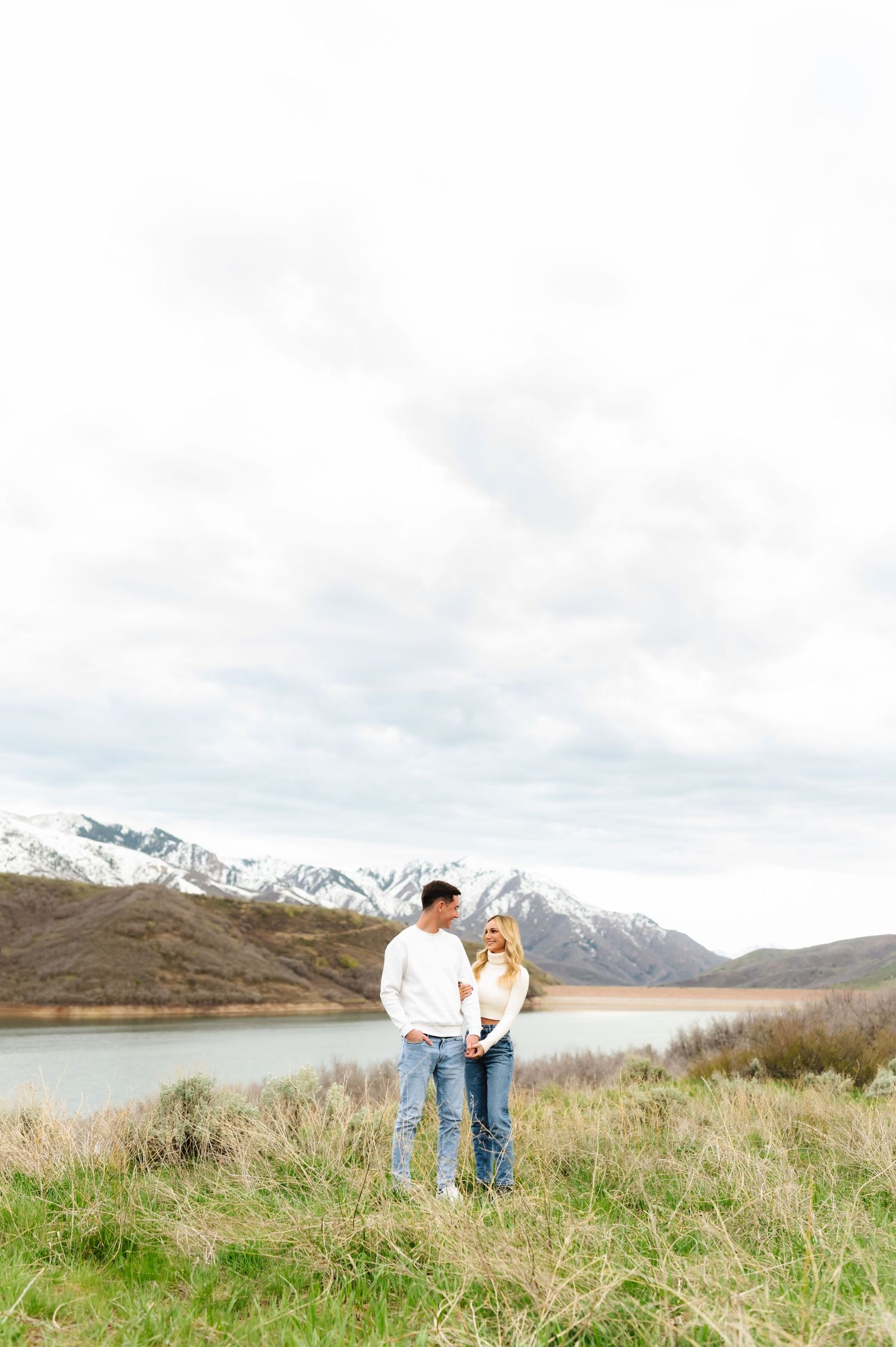 Couple standing on hillside overlooking mountain lake landscape.