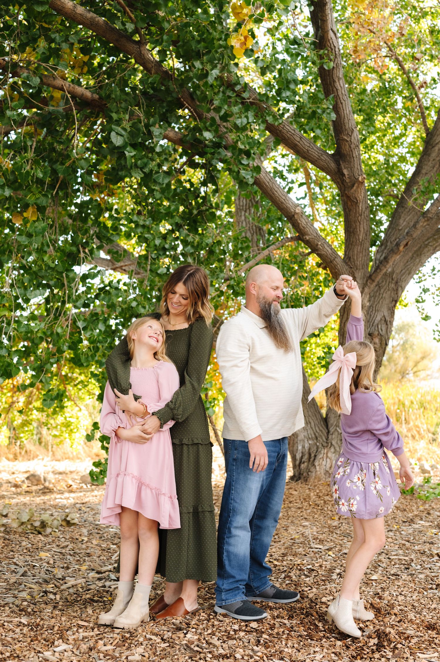 Family of four laughing and playing under a large green tree in autumn, wearing coordinated outfits in pink, olive, and white.