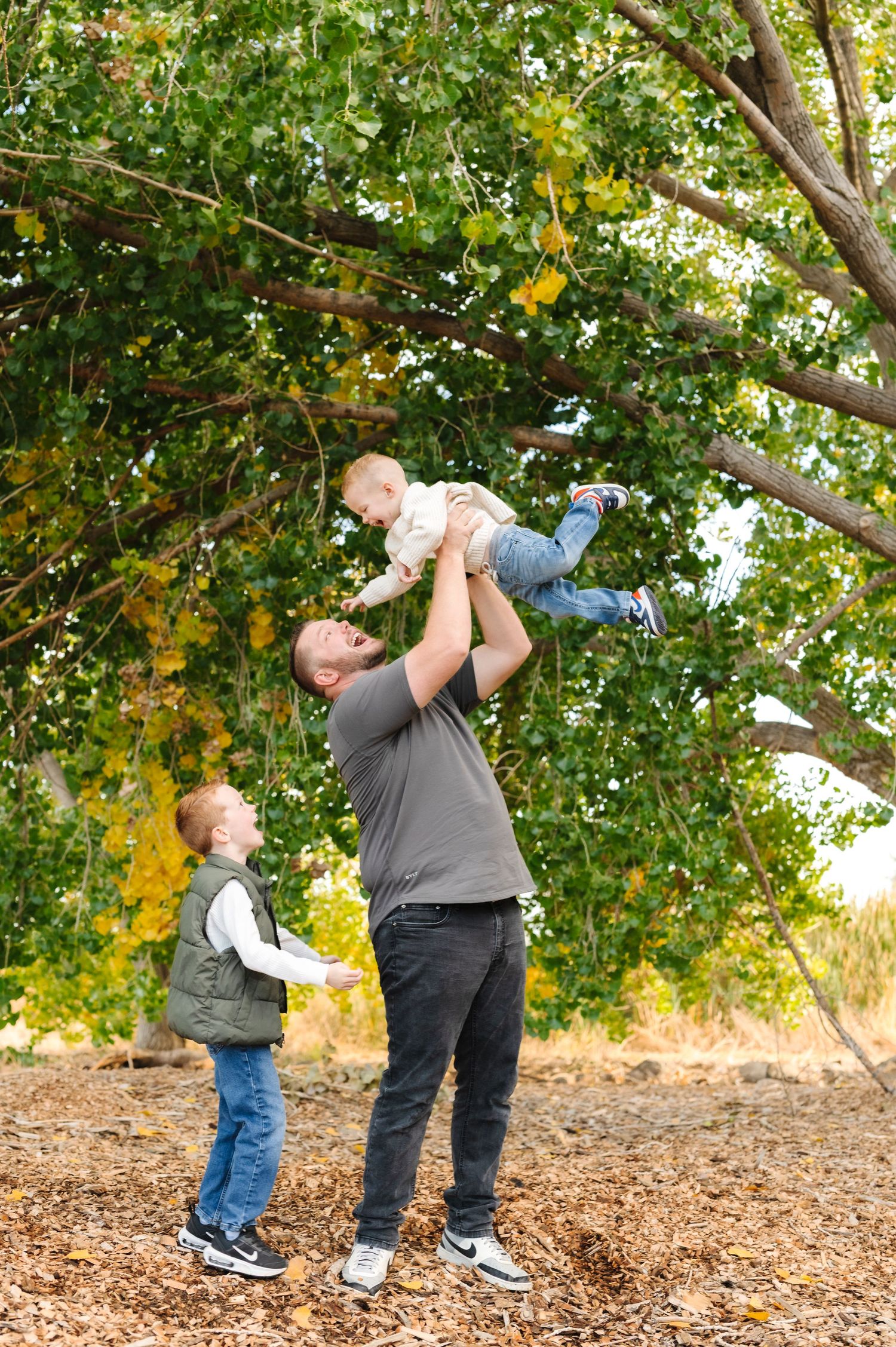 A father lifts a baby in the air outdoors in autumn while a young boy watches, surrounded by colorful fall foliage.