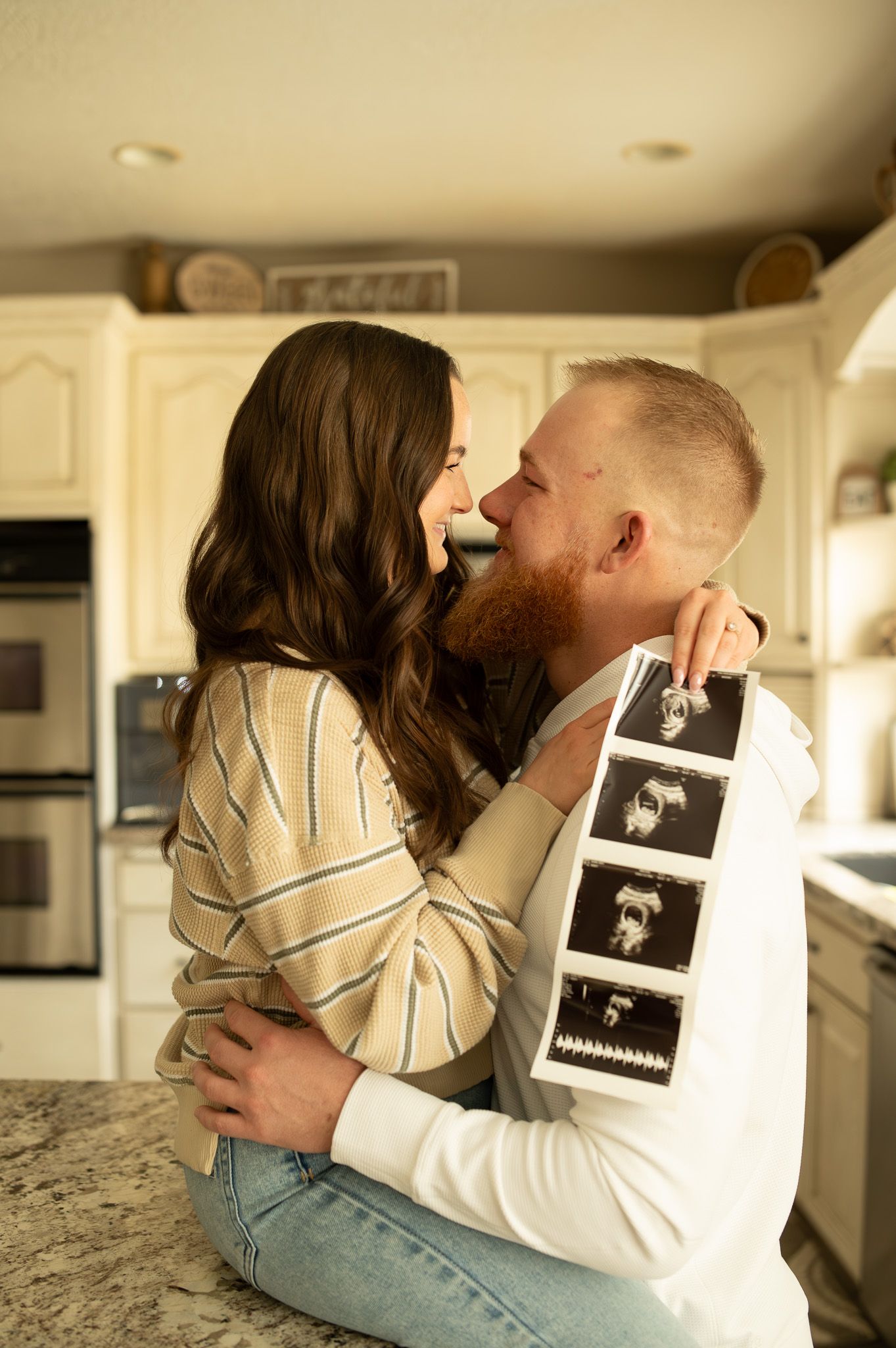 Couple embracing in kitchen holding ultrasound photos, sharing a tender moment for a pregnancy announcement.