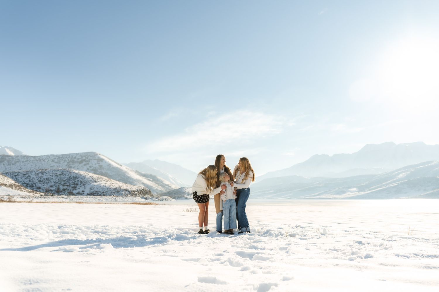 Three women stand together on a snowy frozen lake surrounded by snow-capped mountains on a sunny winter day.