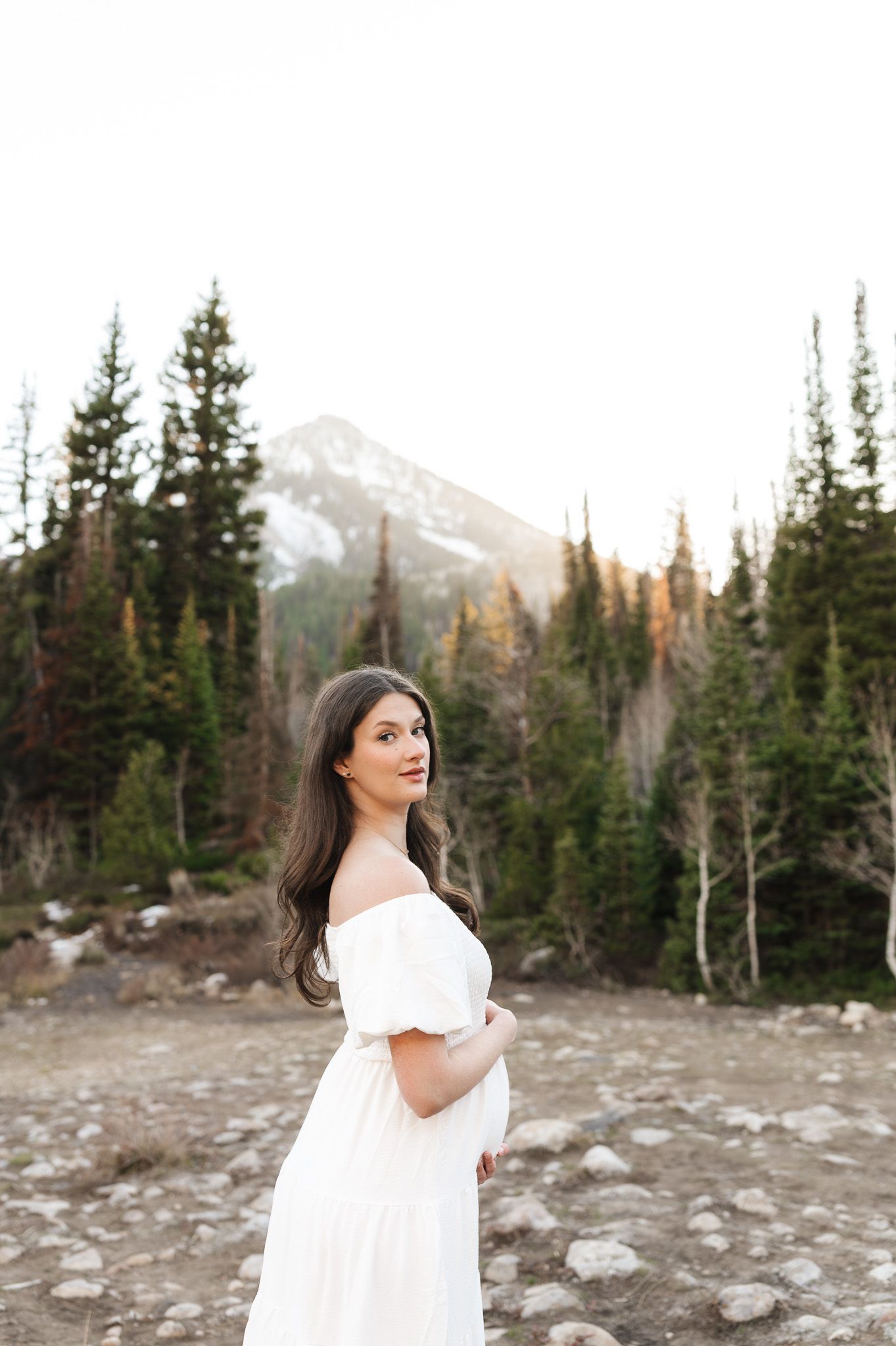 Woman in white off-shoulder dress poses in mountain forest landscape with snow-capped peak backdrop.