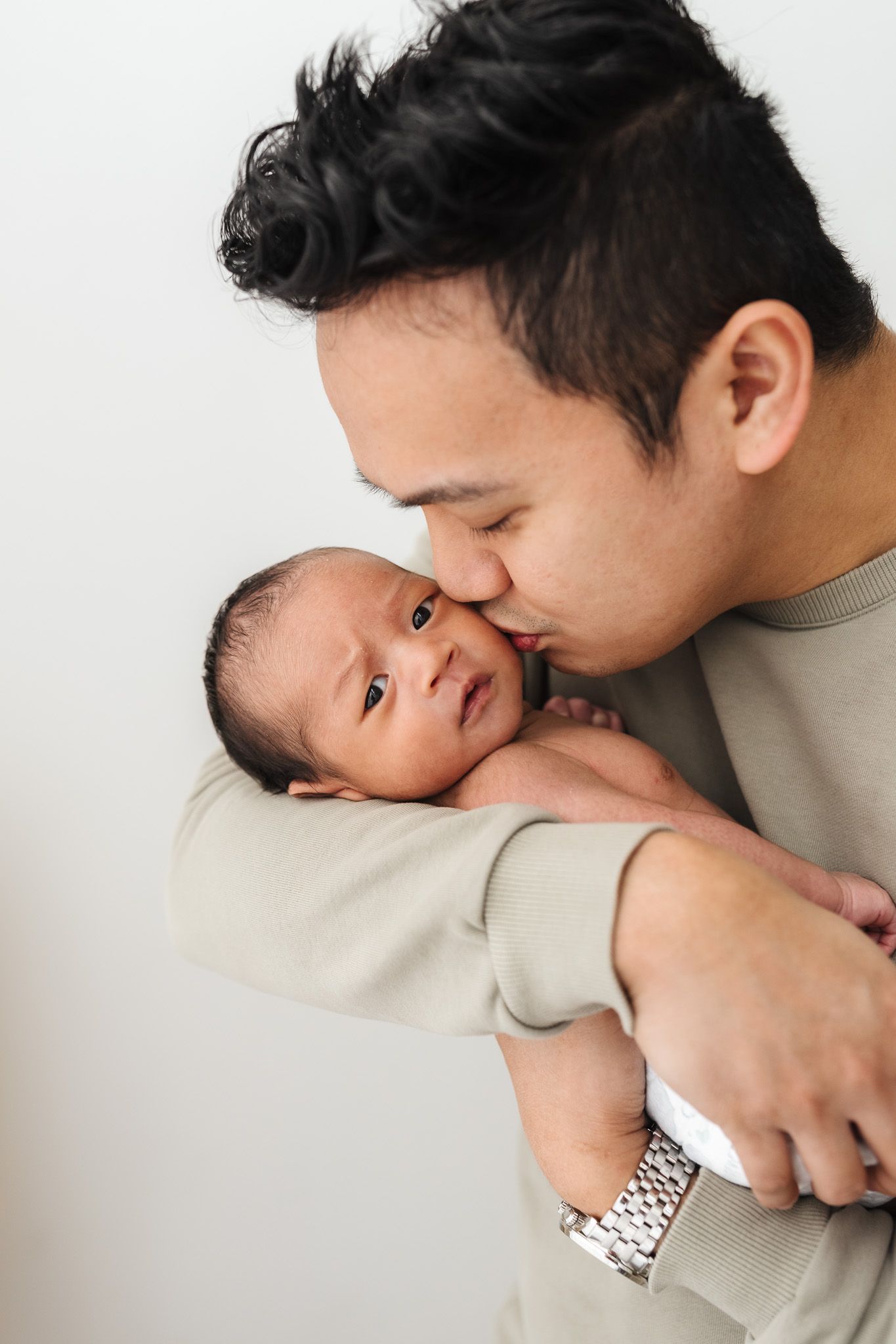 A father tenderly kisses his newborn baby while cradling the infant in his arms against a white background.