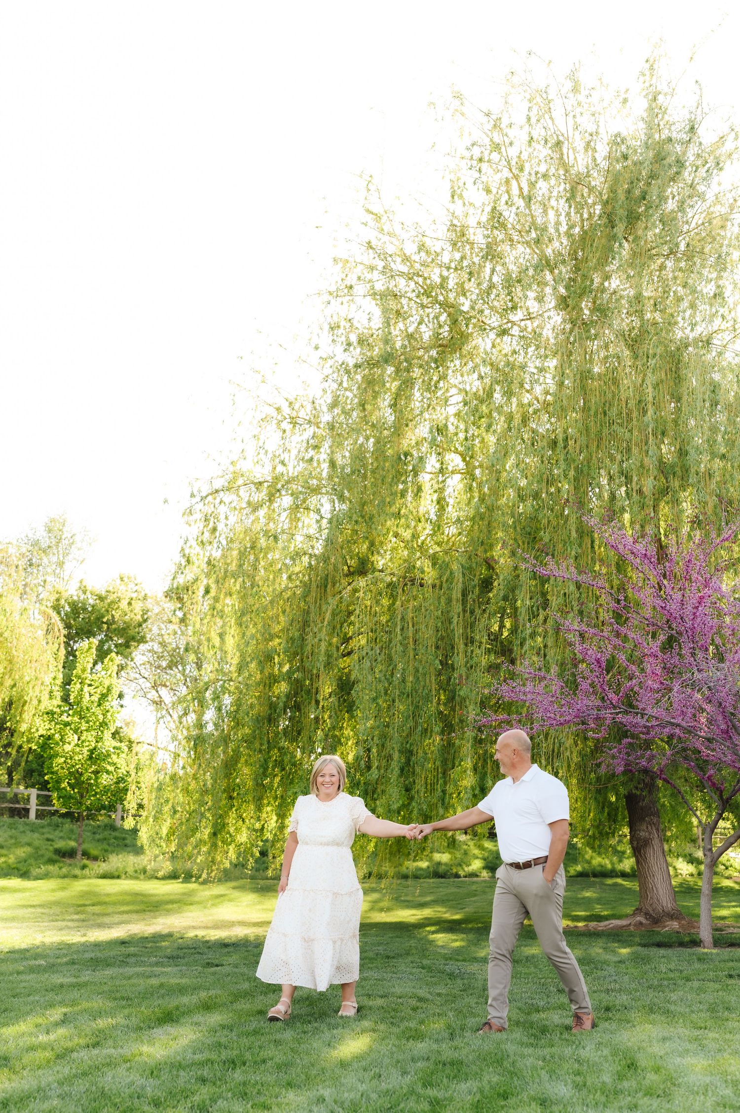 A couple poses together in a garden setting with weeping willow trees and blooming pink flowers in spring.