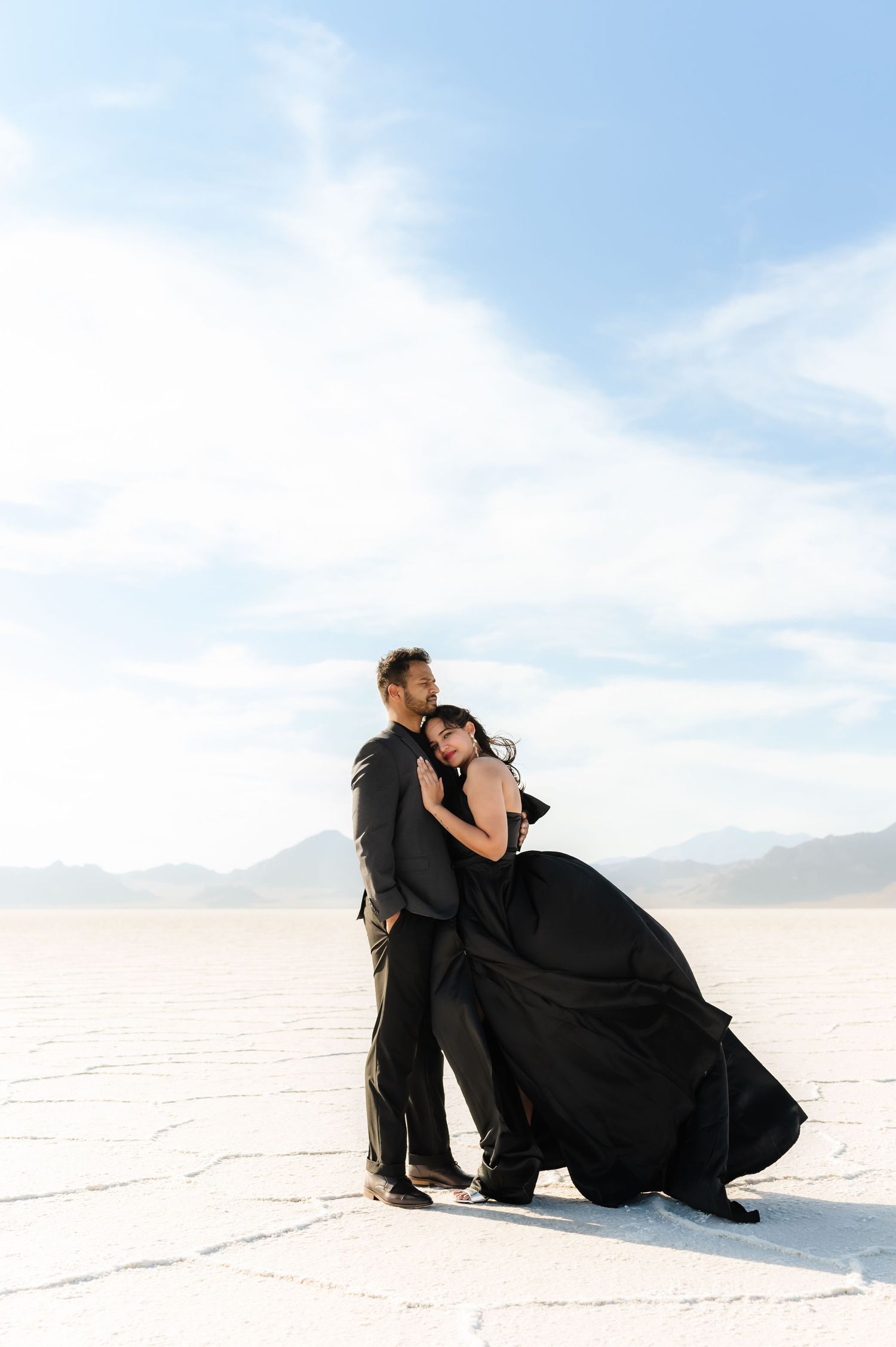 Couple in black formal wear embracing on white salt flats under cloudy sky.