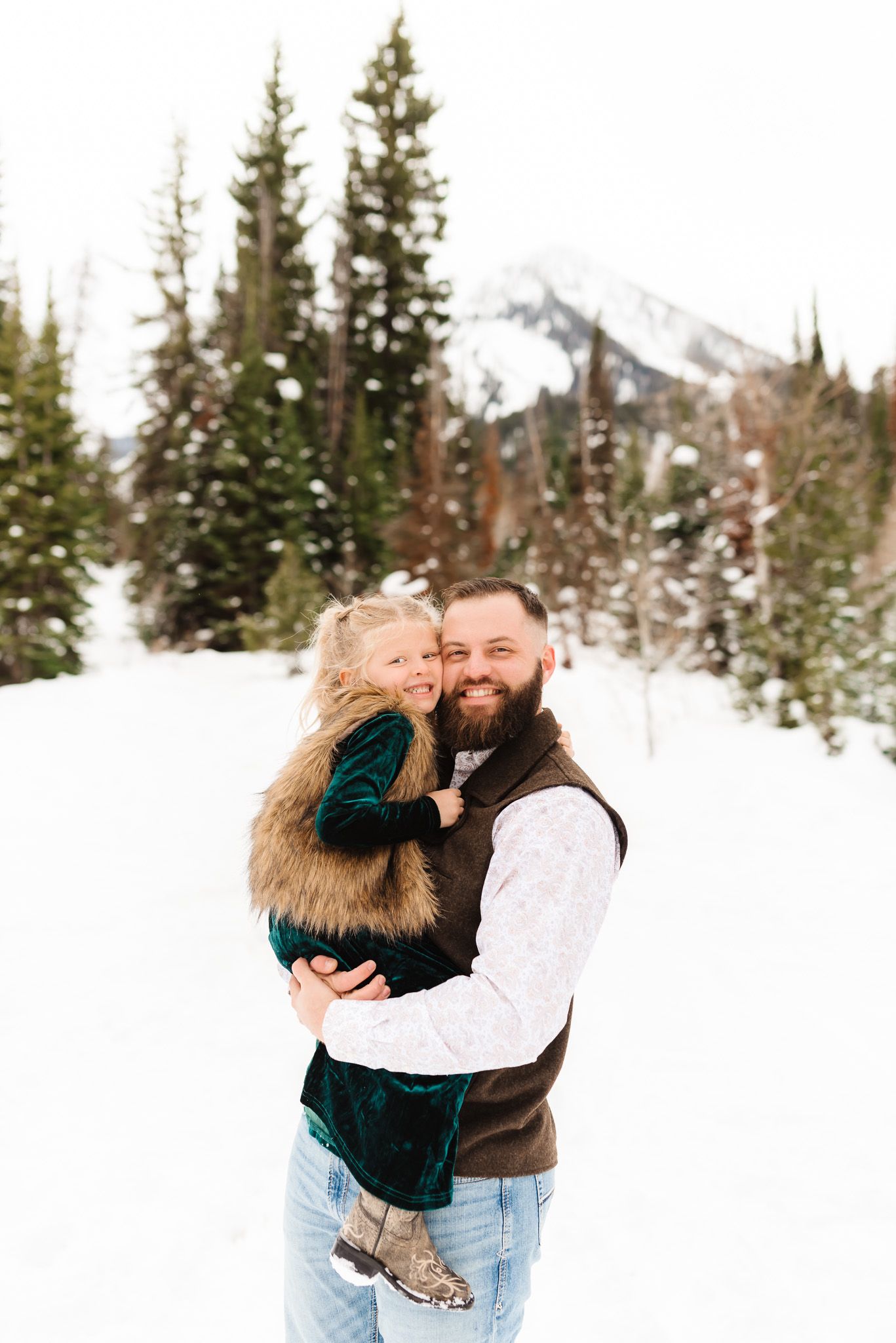 Family embracing in a snowy mountain setting surrounded by evergreen trees.