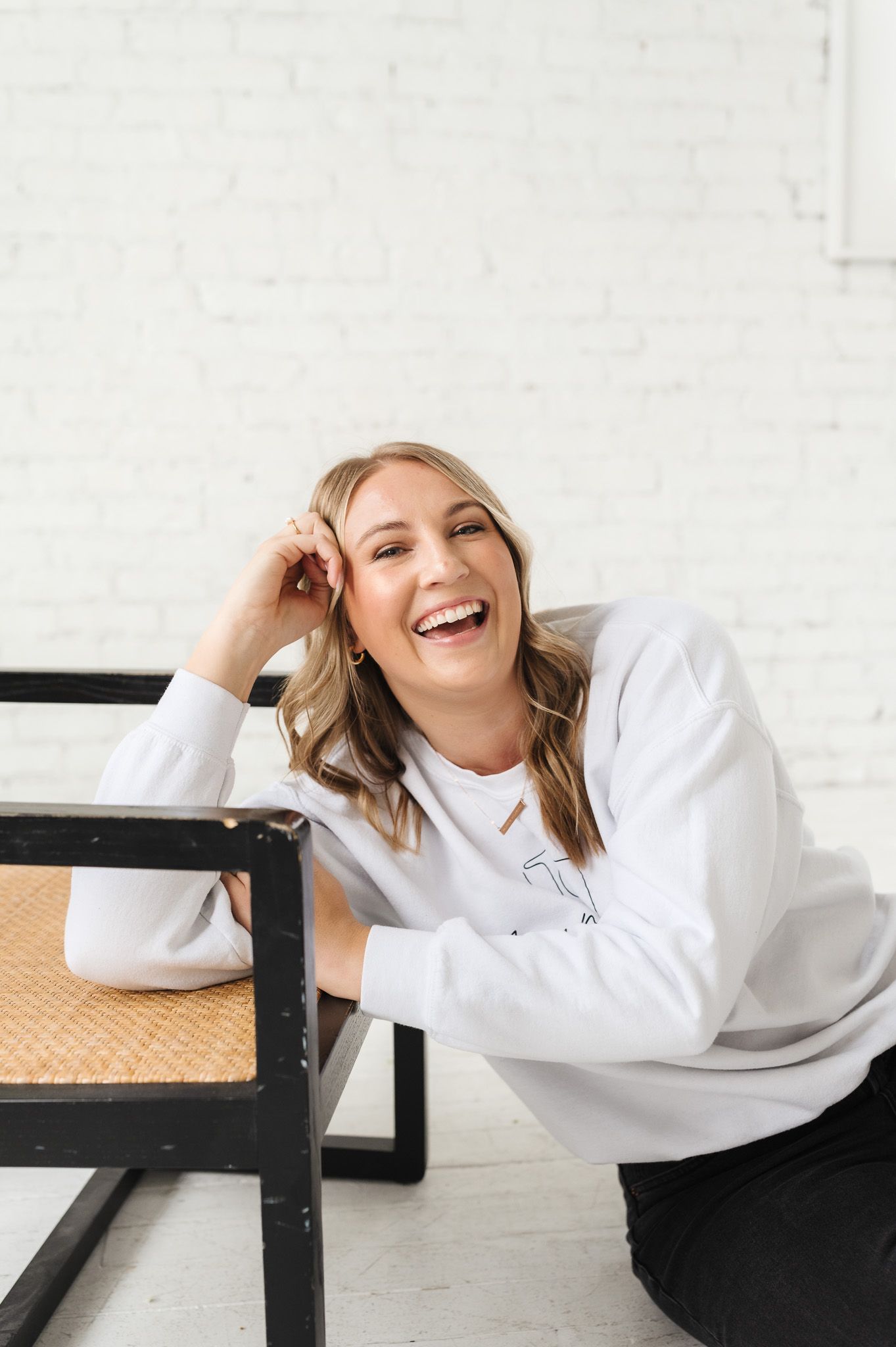 Joyful photographer in white sweater leans on chair in bright studio space.