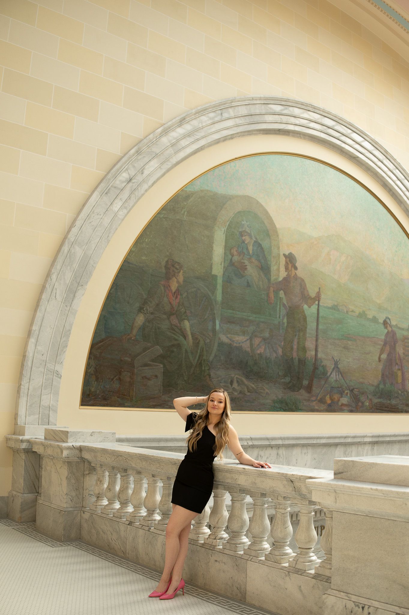 Young woman in black dress poses by ornate balustrade beneath a large classical mural in an elegant neoclassical building.
