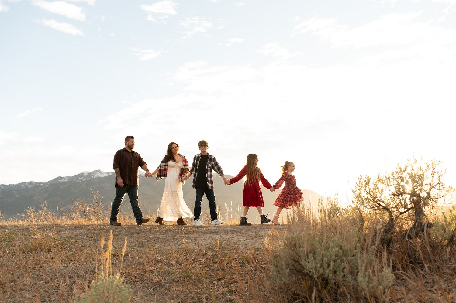 A family of five holds hands walking through a golden field with mountain views at sunset.