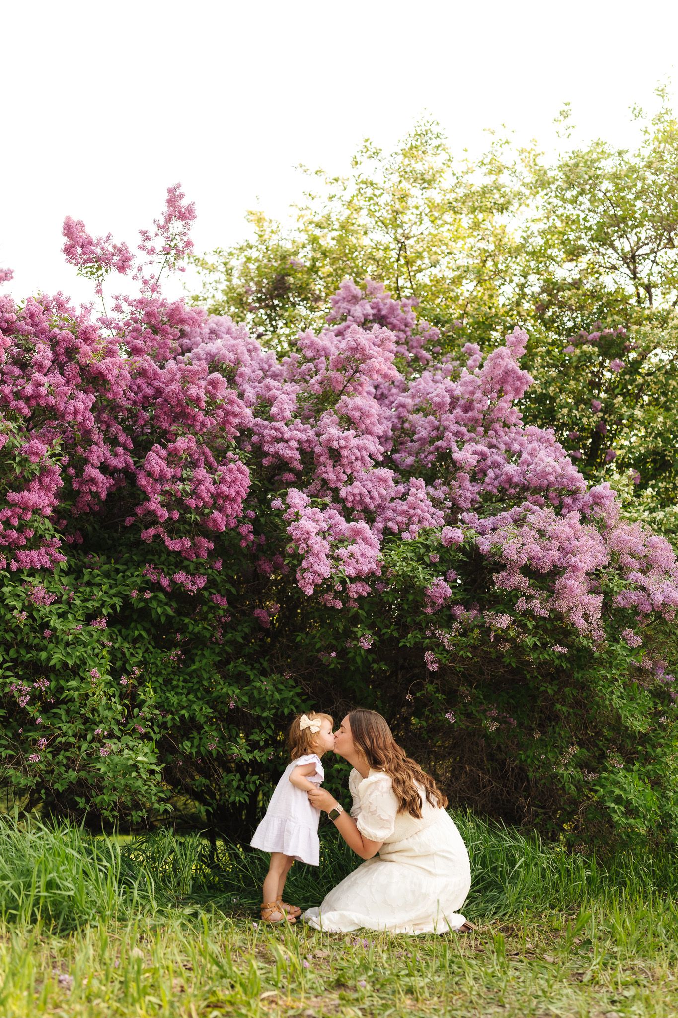 Mother kissing young daughter in white dresses beside blooming purple lilac bushes in a sunny garden.