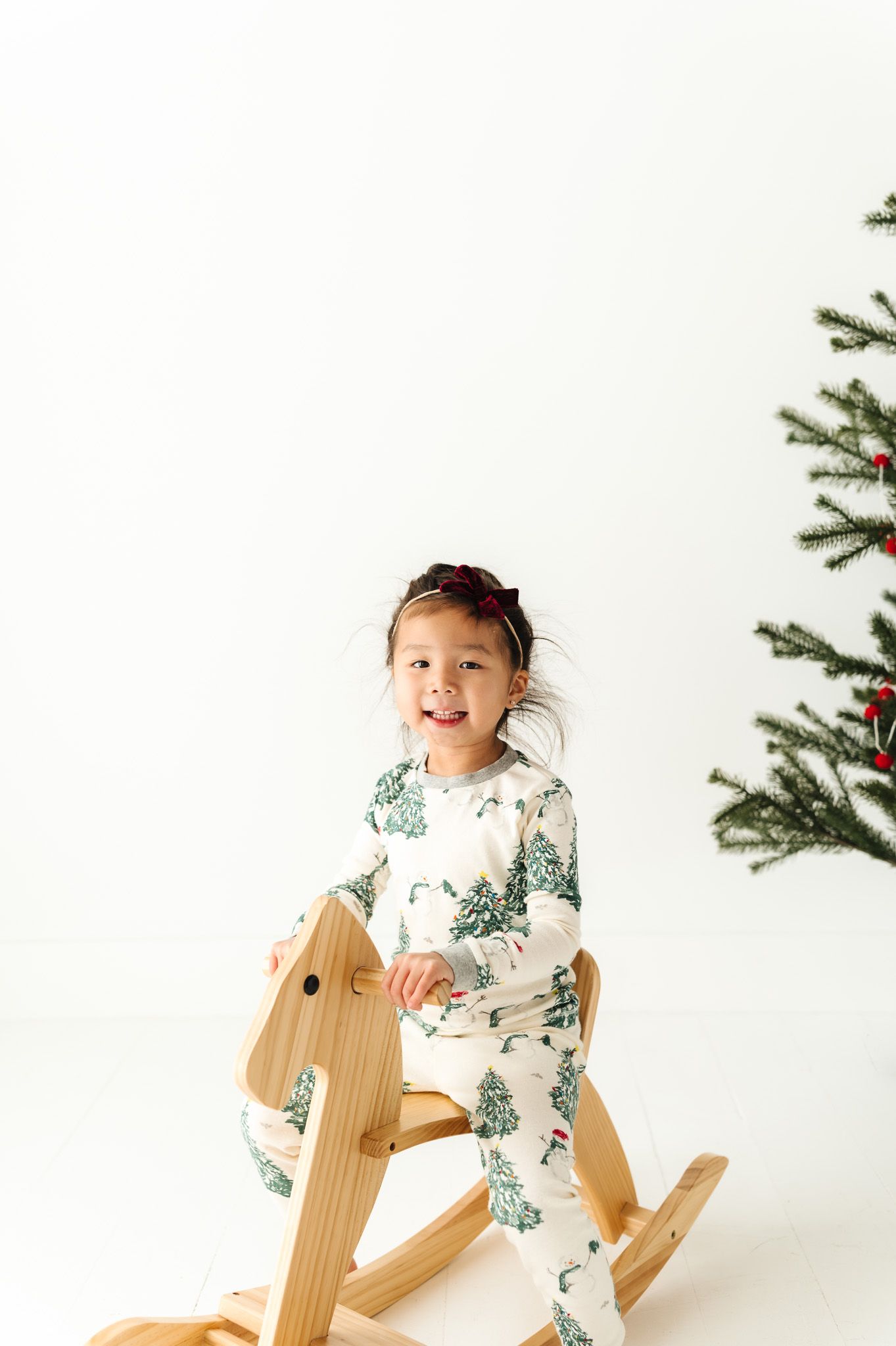 Child in winter-themed pajamas sits on wooden rocking horse next to Christmas tree.