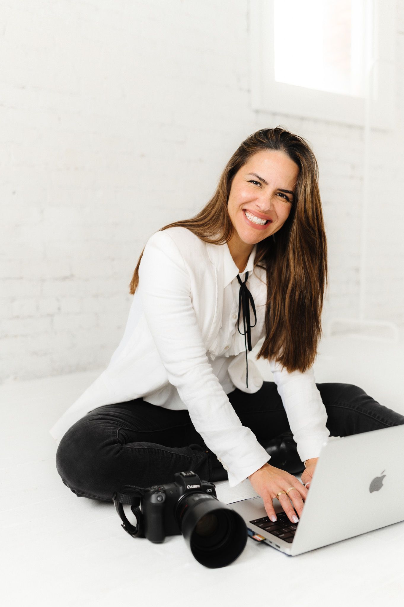 Professional photographer in white blouse works on laptop with camera equipment against white brick wall.