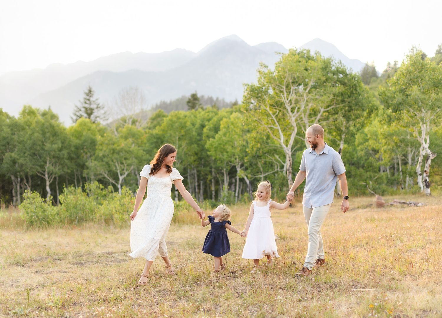 A family walks through a golden meadow with mountains and trees in the background during a sunny end of summer day in American Fork Canyon.