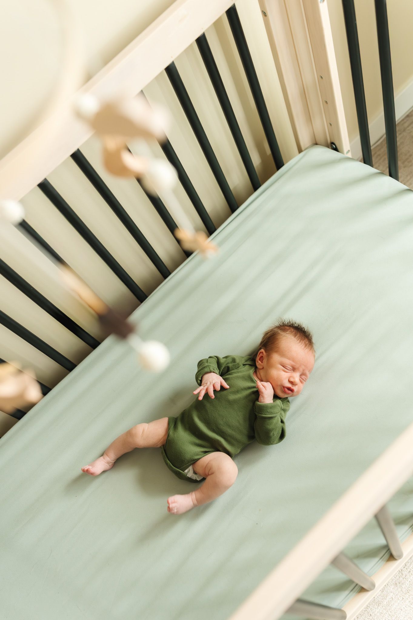 Baby in green onesie lying on mint colored crib mattress seen through wooden crib slats.