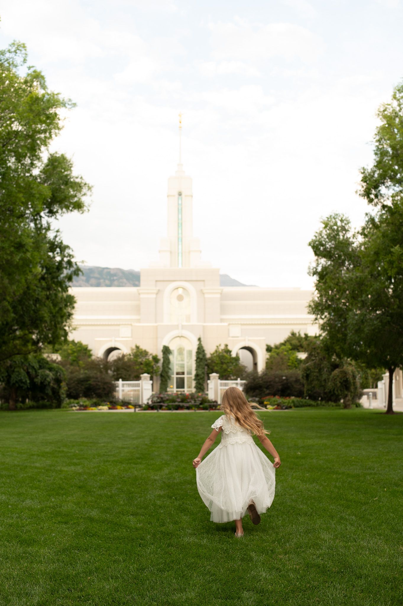 Young girl in white dress running toward an LDS temple on a green lawn with trees and mountains in background.