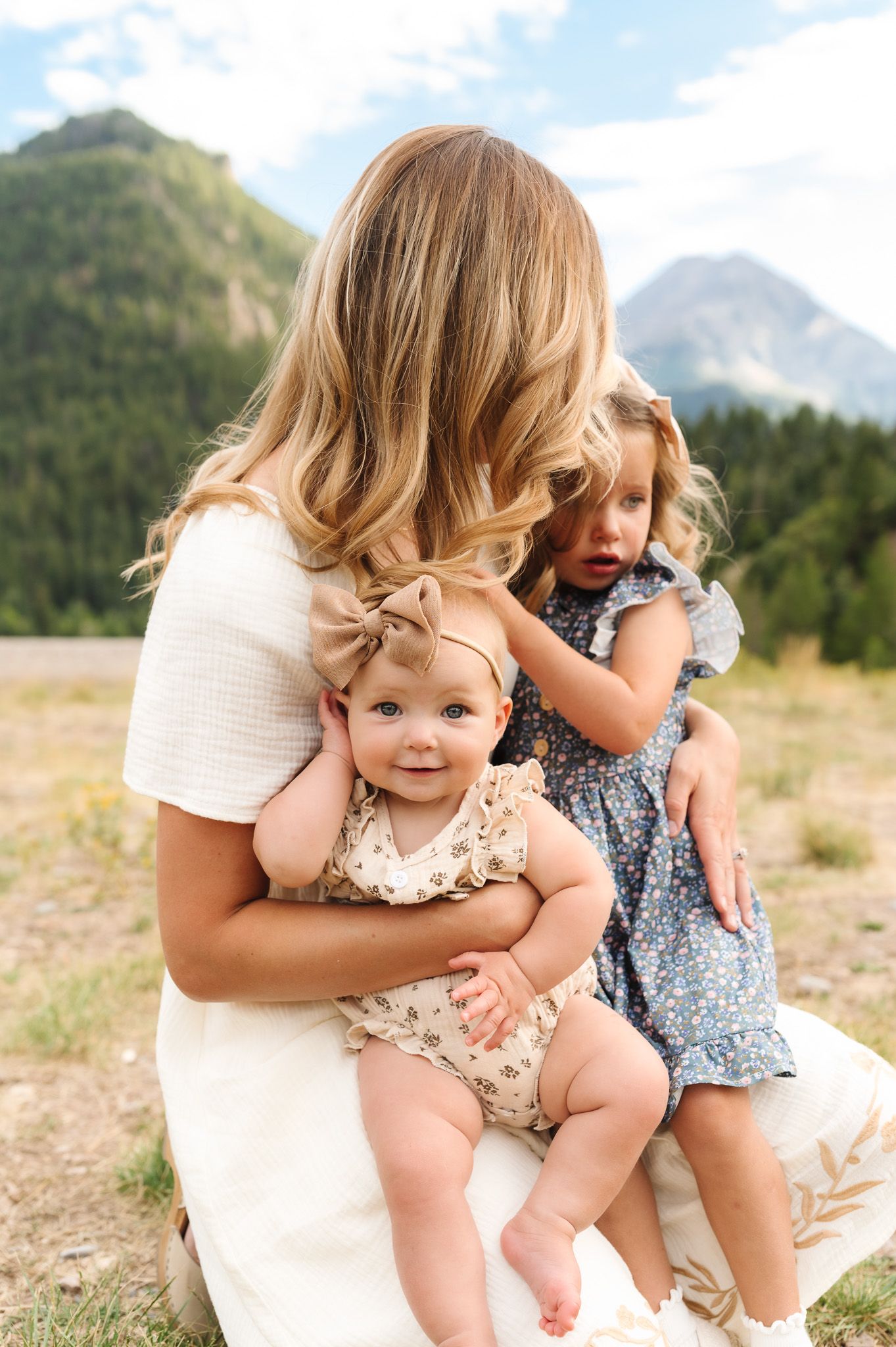A mother holds her baby and toddler daughter outdoors with mountains in the background.