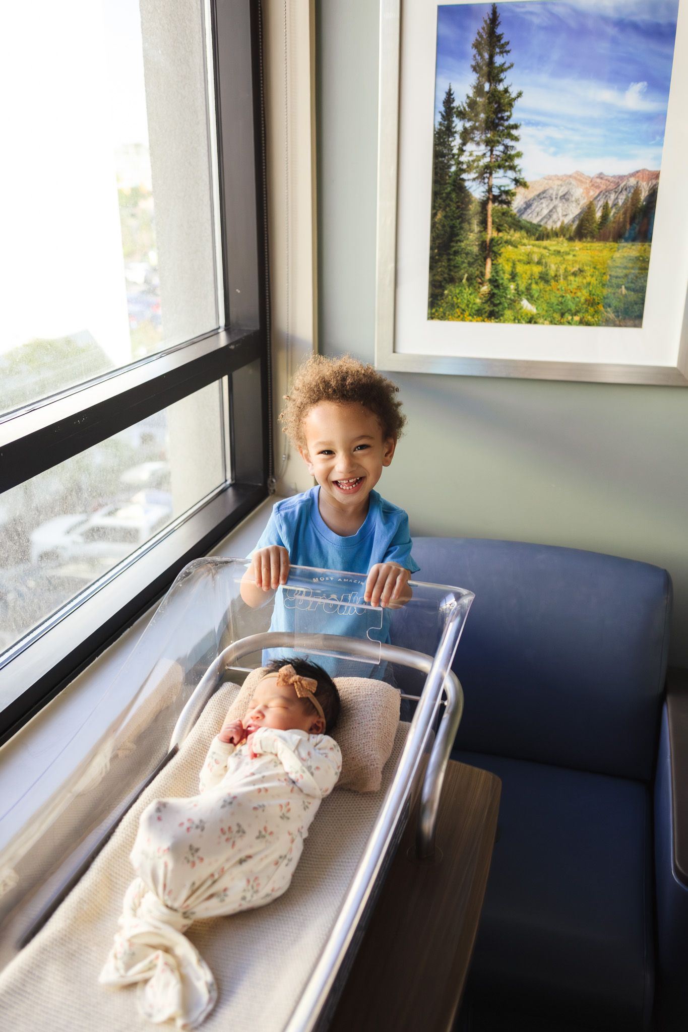 Smiling child stands beside hospital bassinet with swaddled newborn baby near window in maternity ward.