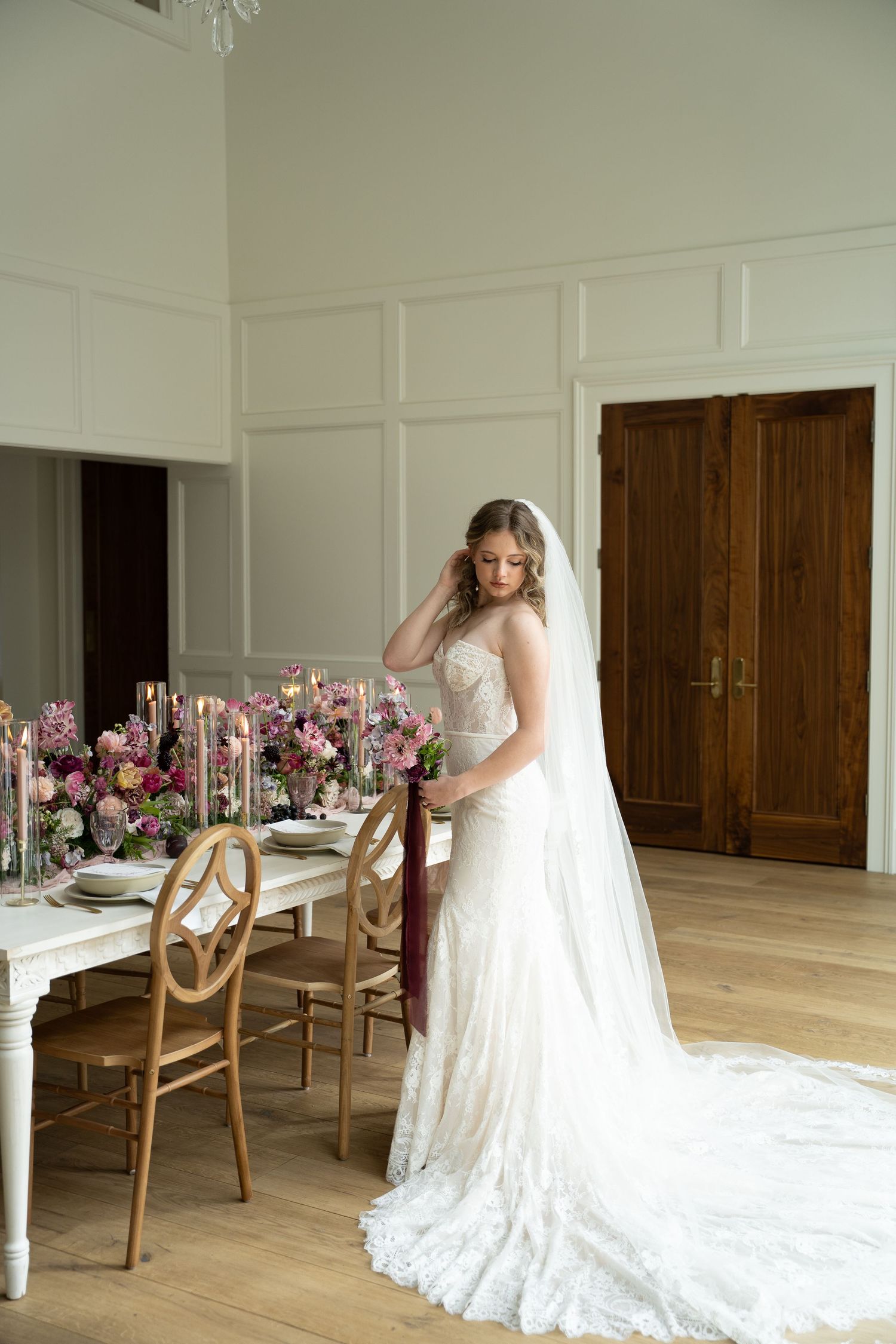 Elegant wedding dress with long train displayed in bright white room with wooden chairs and floral table arrangements.
