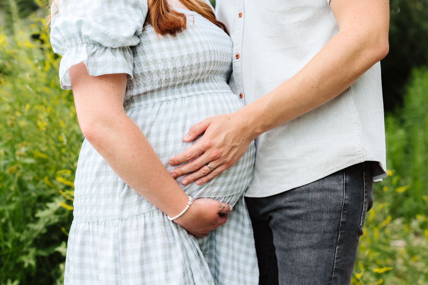 Close up of hands cradling a pregnant belly in a blue gingham dress outdoors.
