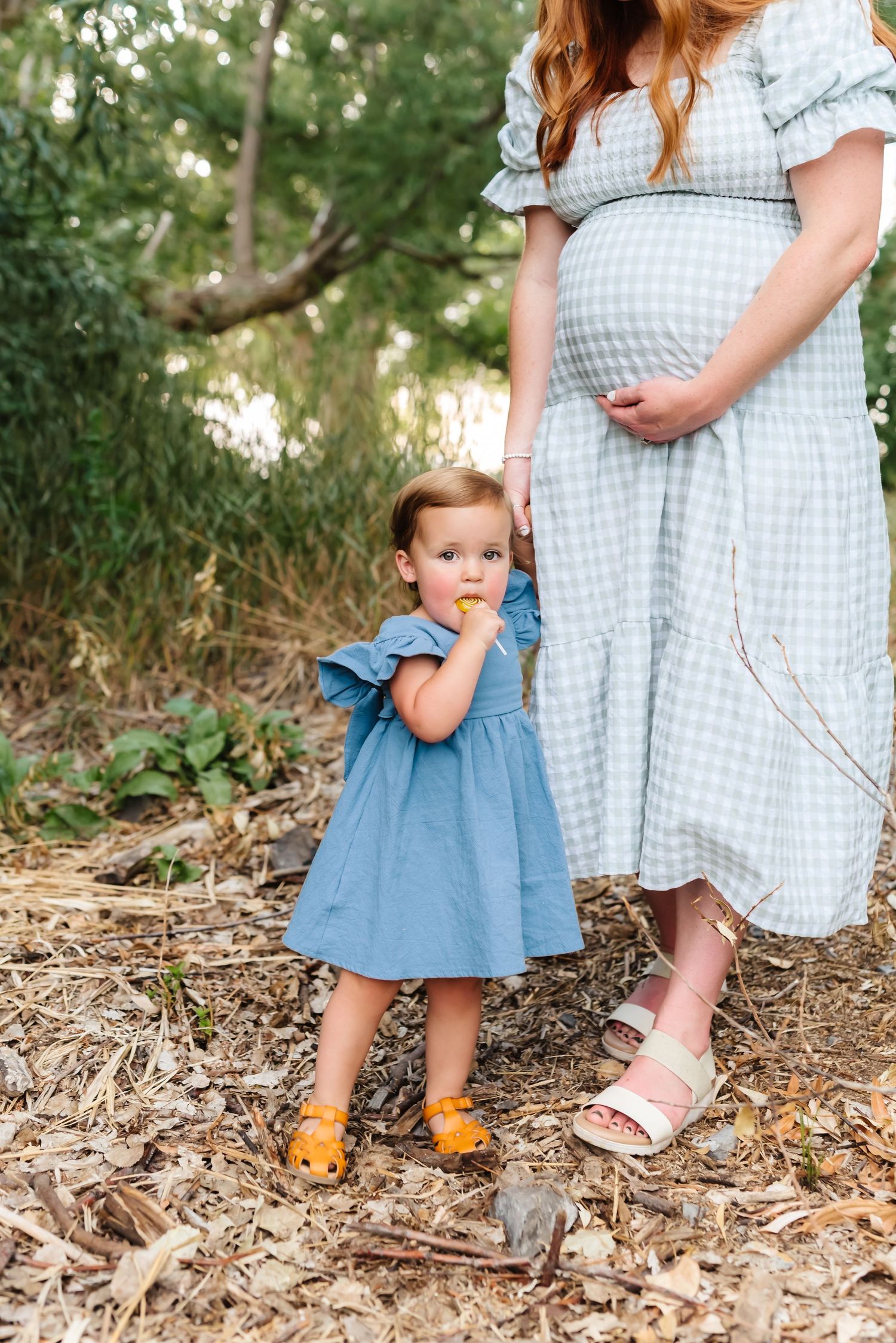Mother in gingham dress and child in blue dress standing together on woodland ground.