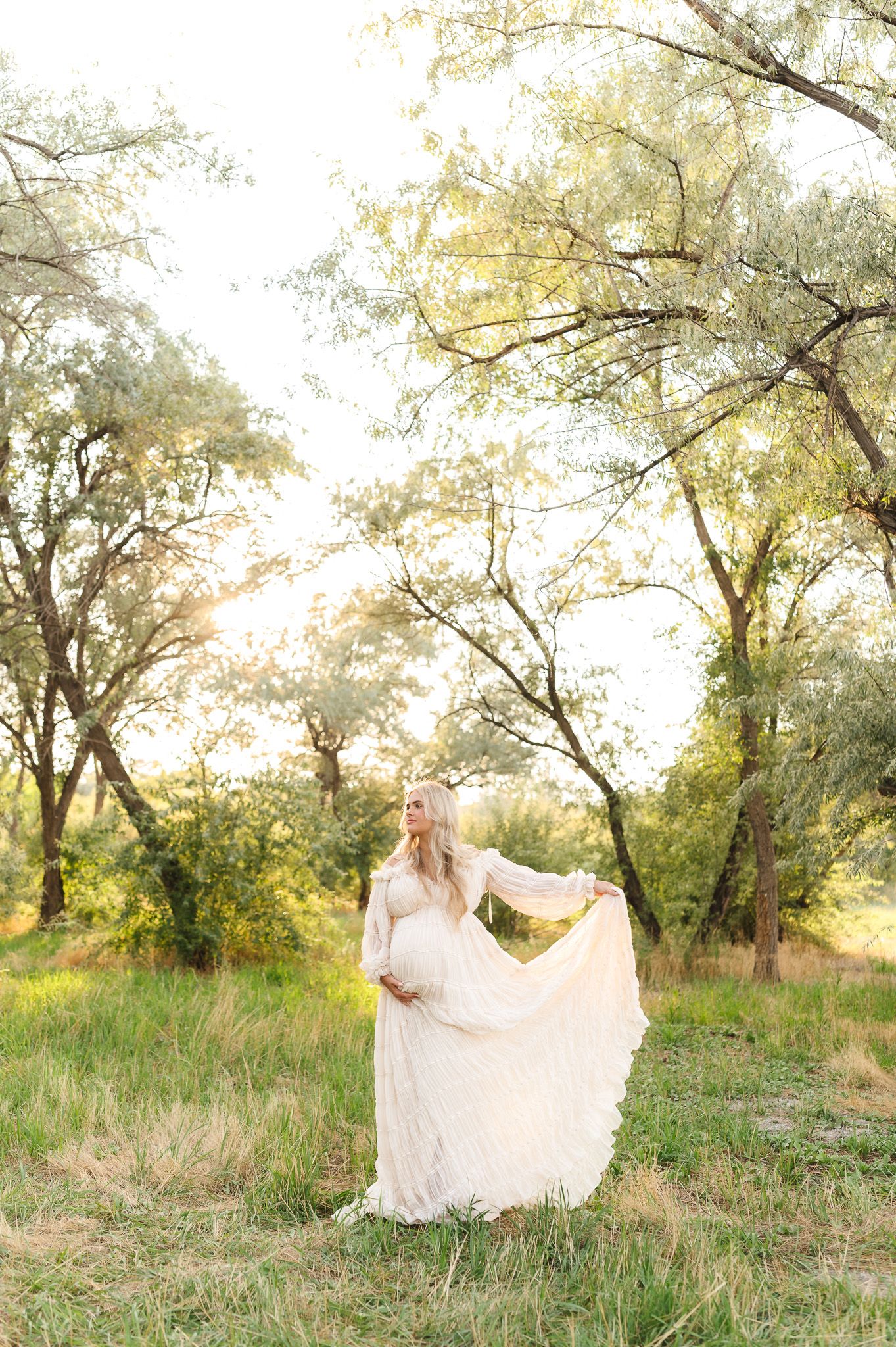 Pregnant woman in flowing white lace gown holding dress in sunlit meadow surrounded by trees.