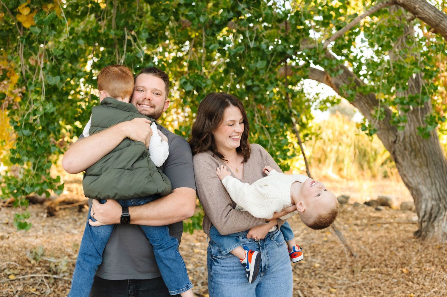 Happy family with two young children laughing outdoors under a leafy tree during a fall photo session.
