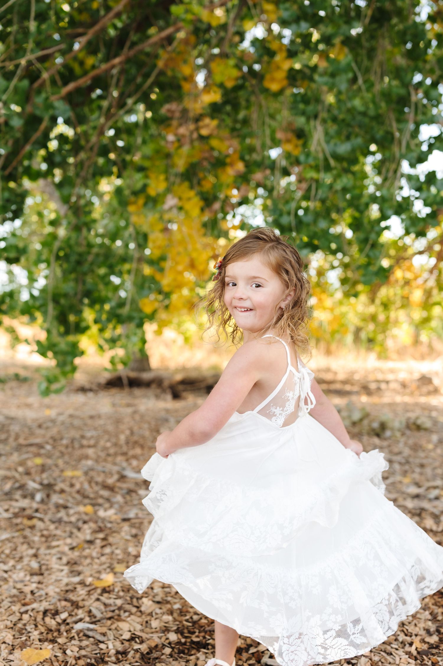 A young girl in a white lace dress twirls outdoors under green and yellow trees on a sunny day.