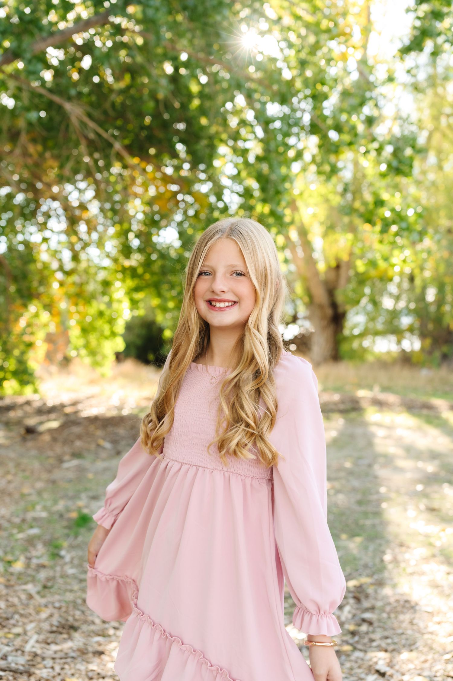 Smiling young girl with long blonde hair wearing a pink long-sleeve dress, posing outdoors in a sunlit park.