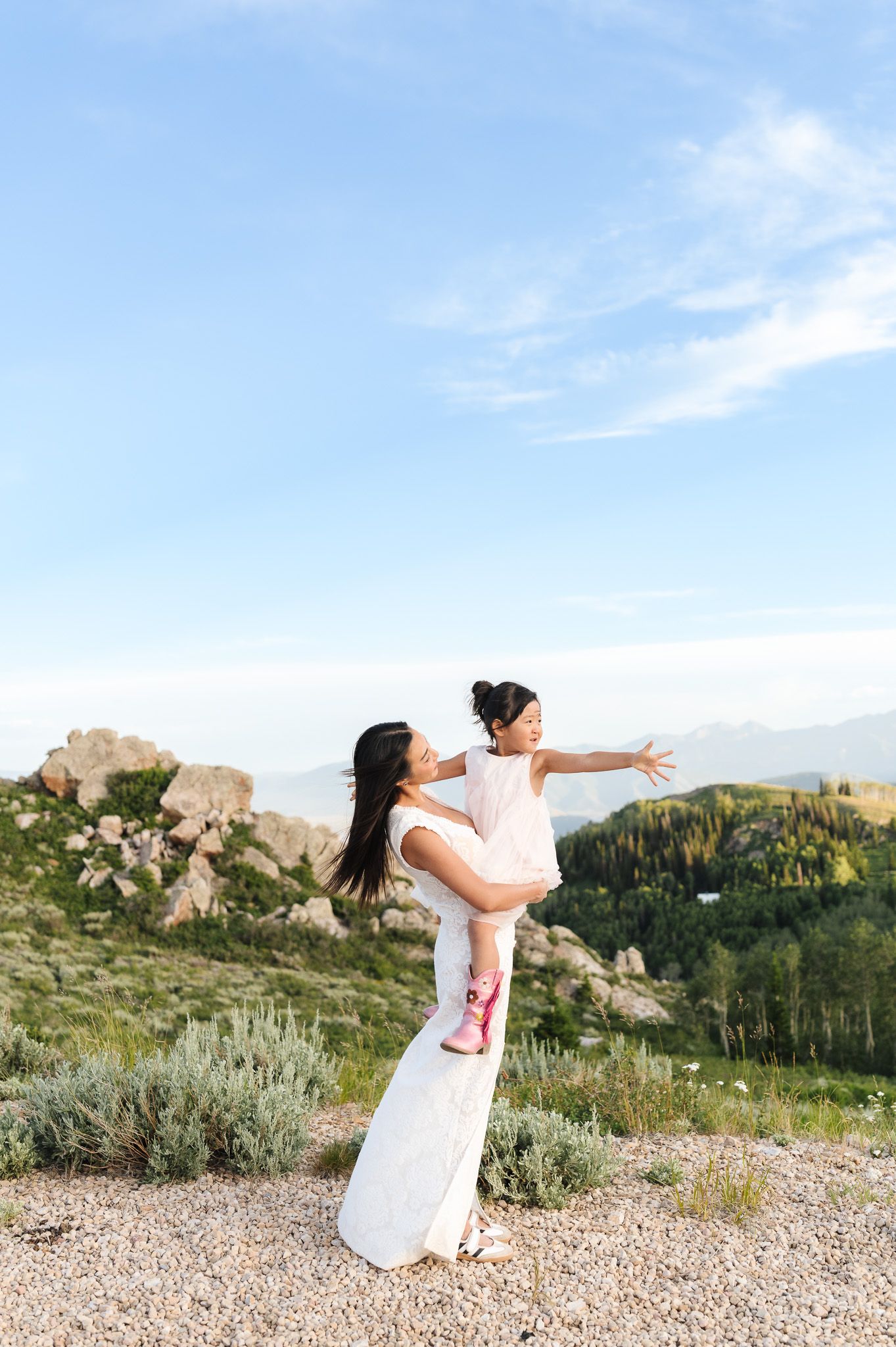A woman in a white dress lifts a young girl with outstretched arms on a scenic mountain overlook.
