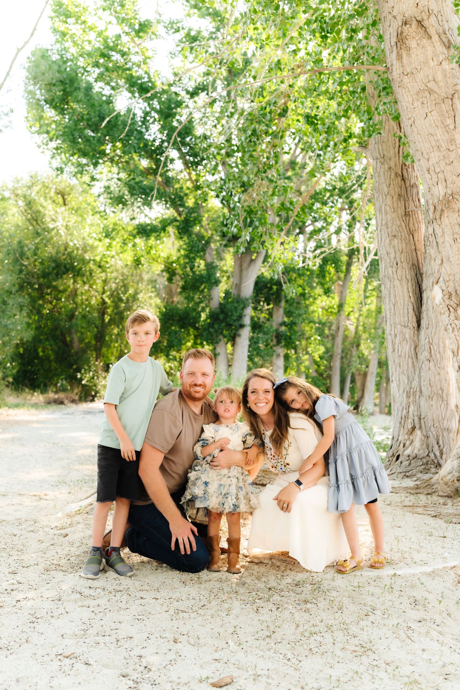 Family group relaxing together on ground near large trees in casual outdoor portrait session.
