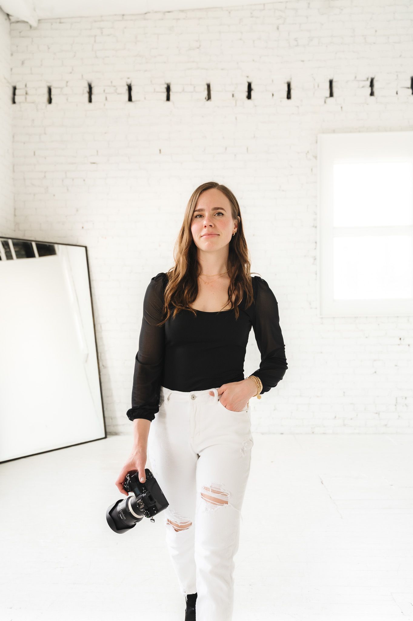 Photographer in black top and white jeans stands confidently in minimalist studio space.