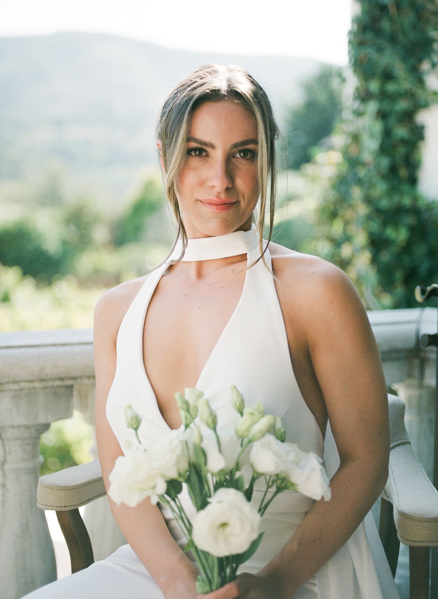 Elegant bride in a white halter dress holds a delicate bouquet of white ranunculus flowers against a dreamy outdoor backdrop.