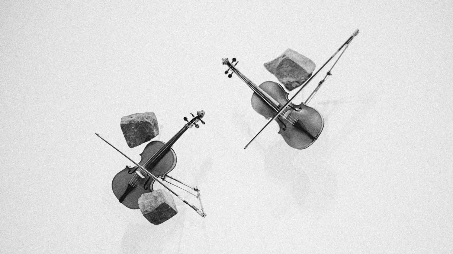 Two cellos with bows resting on rocks, captured in a minimalist black and white aerial photograph.