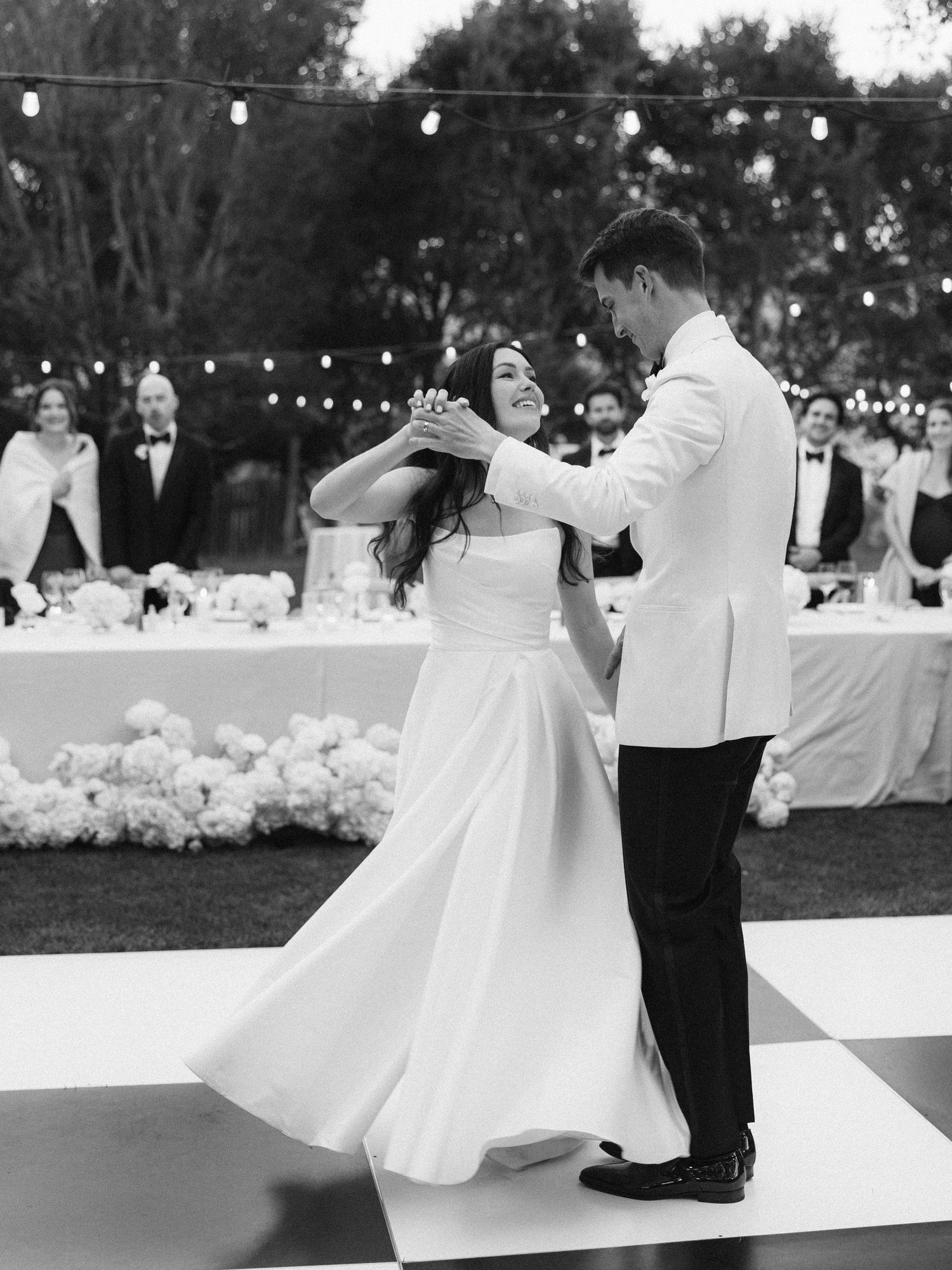 Bride and groom share first dance on checkered floor at elegant outdoor wedding reception with string lights.