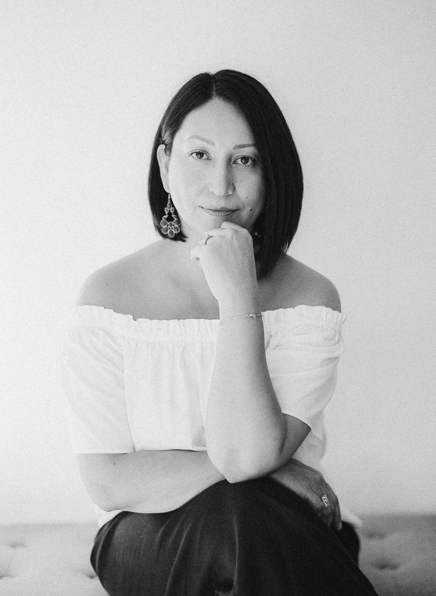 Black and white portrait of a woman in off-shoulder top, posing thoughtfully with hand near chin, wearing ornate earrings.