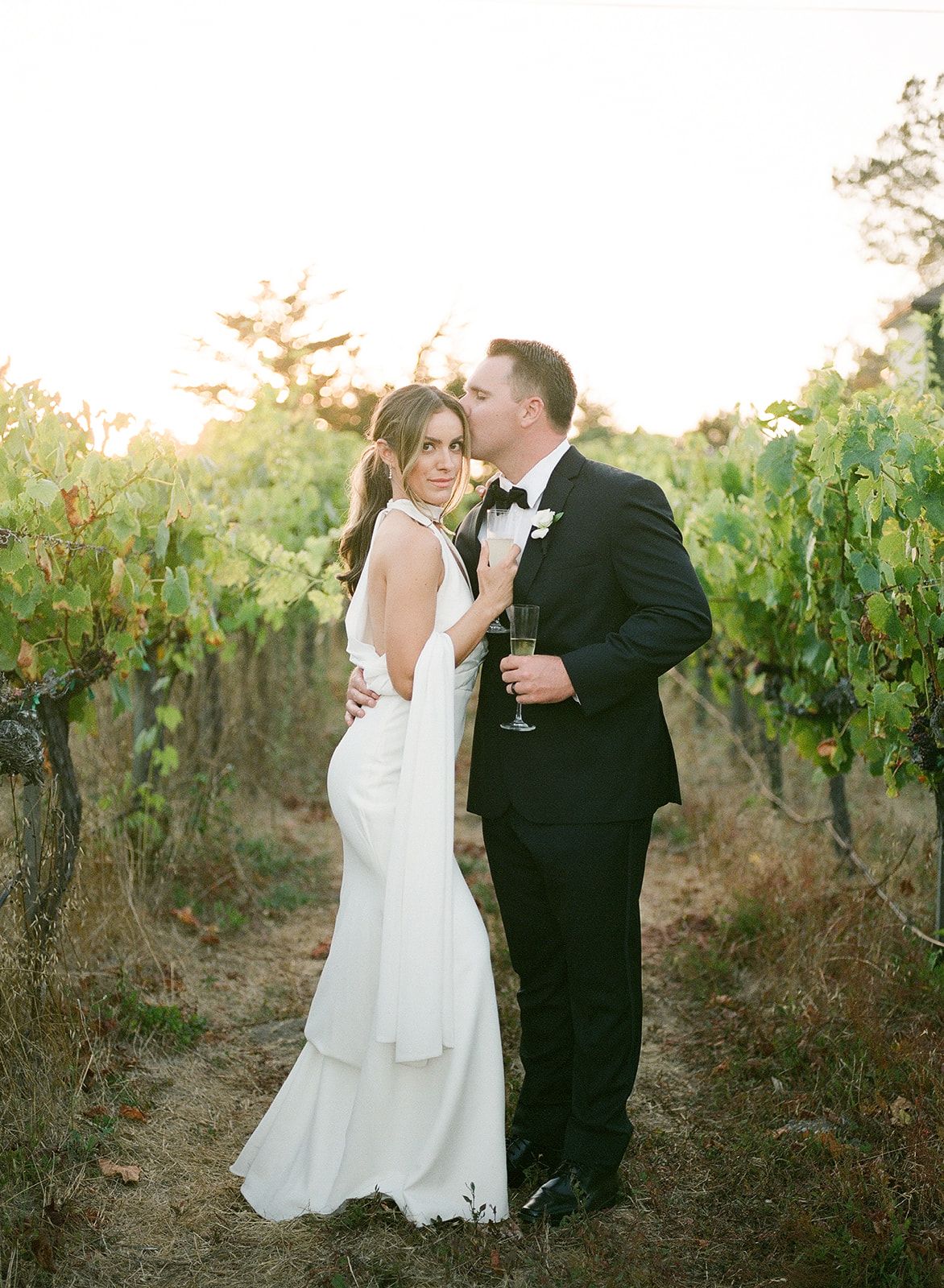 Romantic vineyard wedding photo with a couple enjoying champagne at sunset among the grapevines.