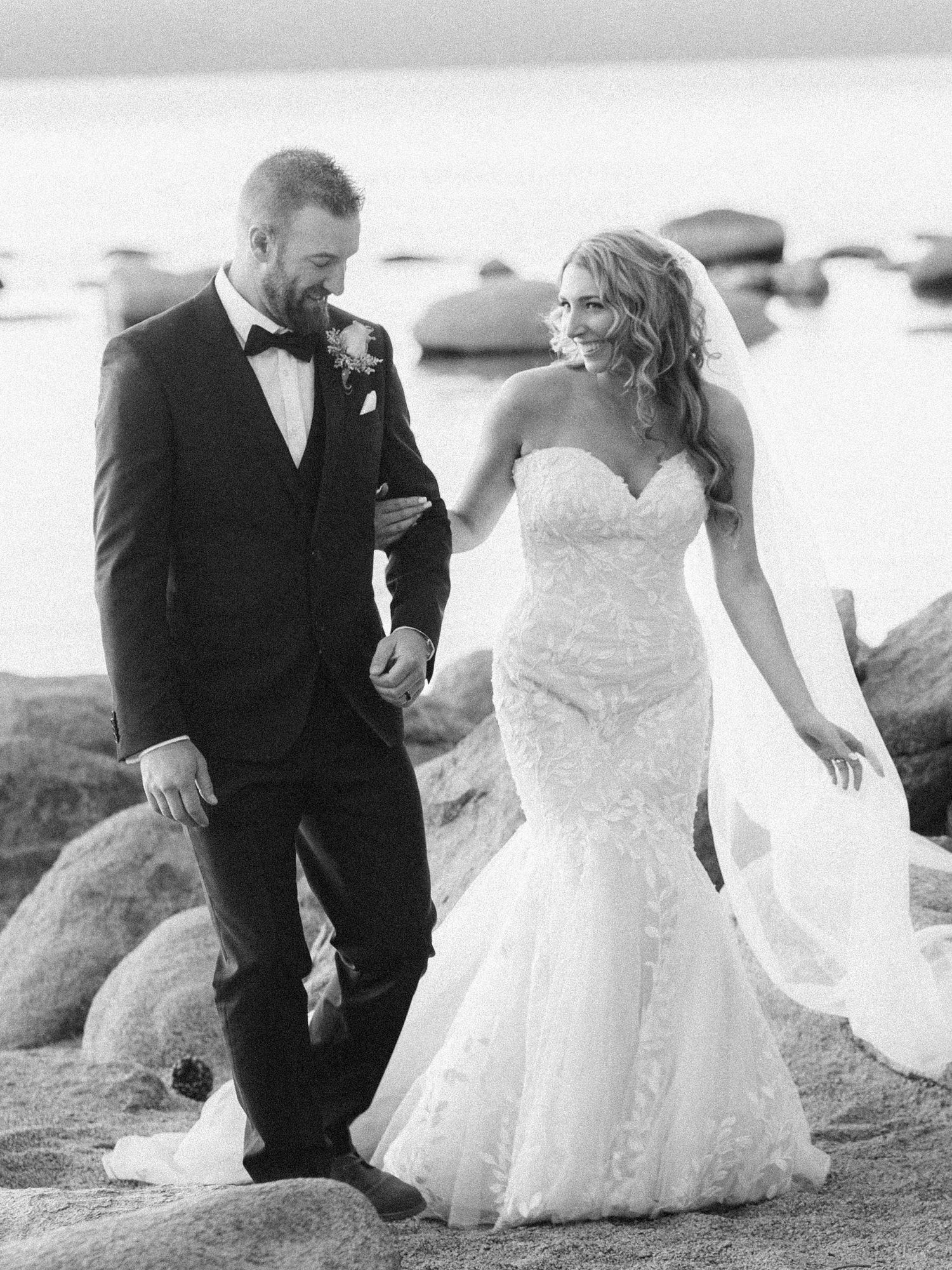 A wedding couple walks together on rocky coastal terrain, with the bride in a lace mermaid gown and groom in black tuxedo.