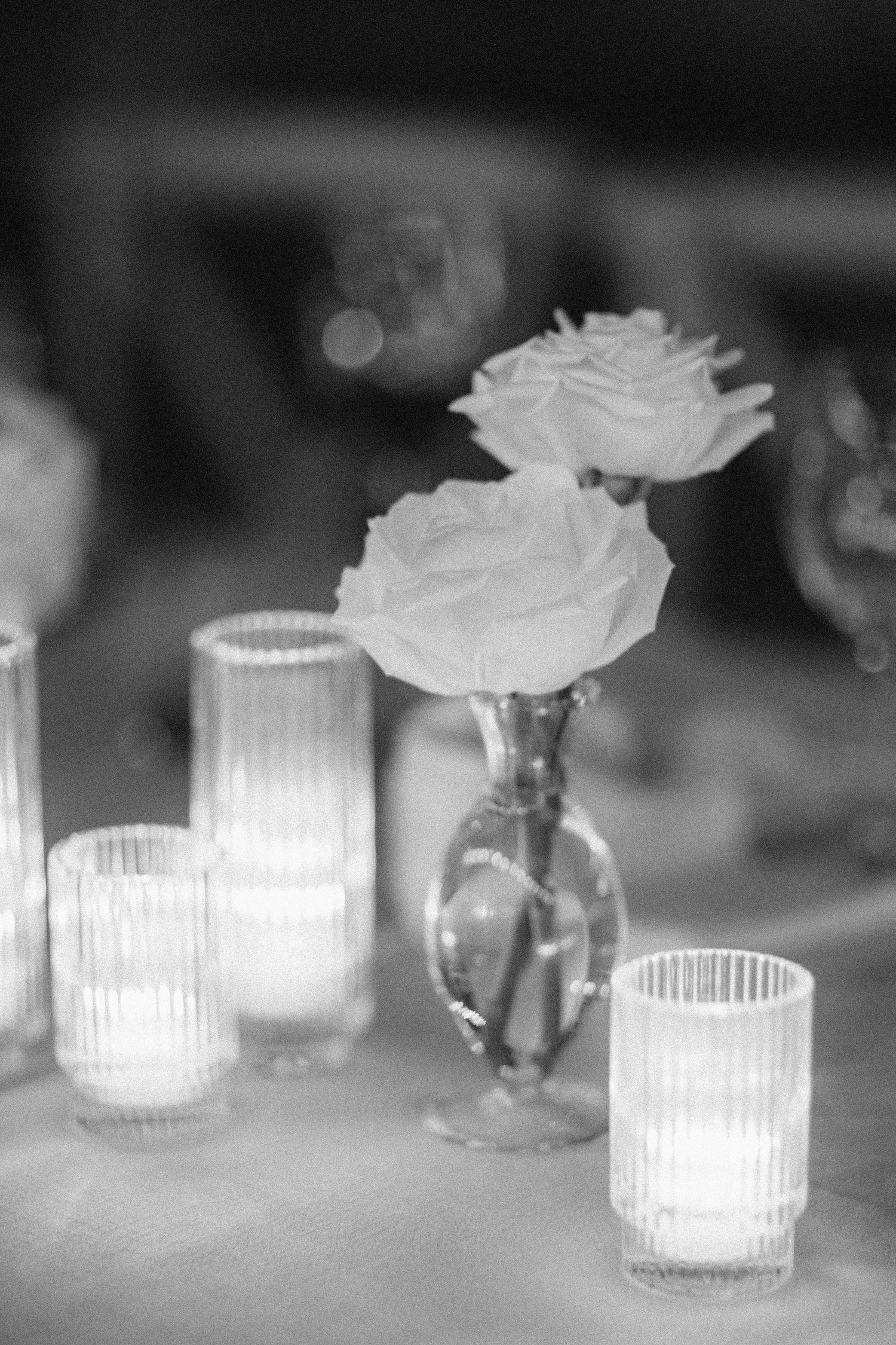 Black and white photo of white roses in a glass vase surrounded by glowing ribbed votive candles on a table.