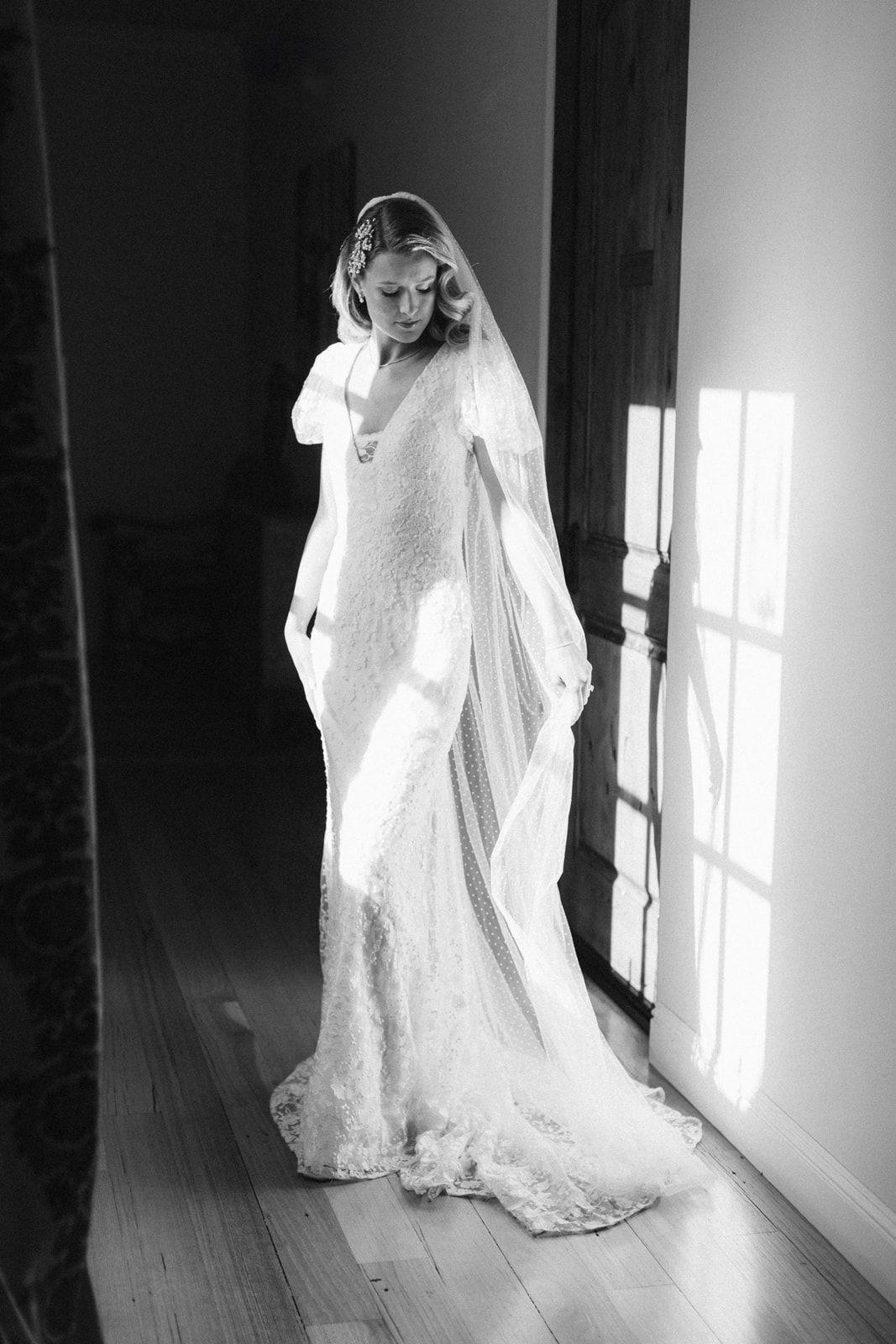 A bride in a white lace wedding gown with long veil stands gracefully by sunlit windows creating a dramatic silhouette.