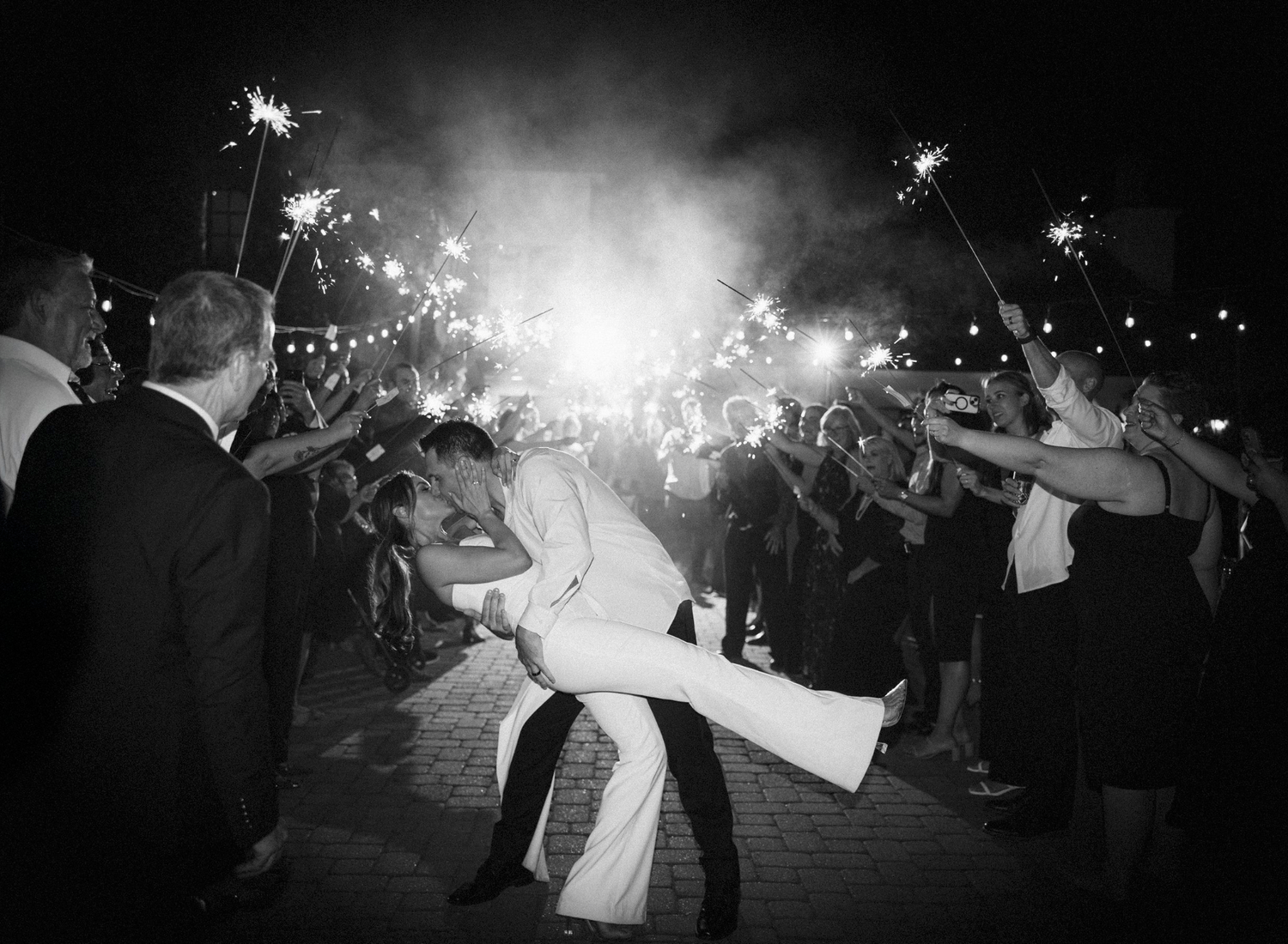 A black and white photograph of a wedding celebration with sparklers illuminating a romantic dance dip at night.