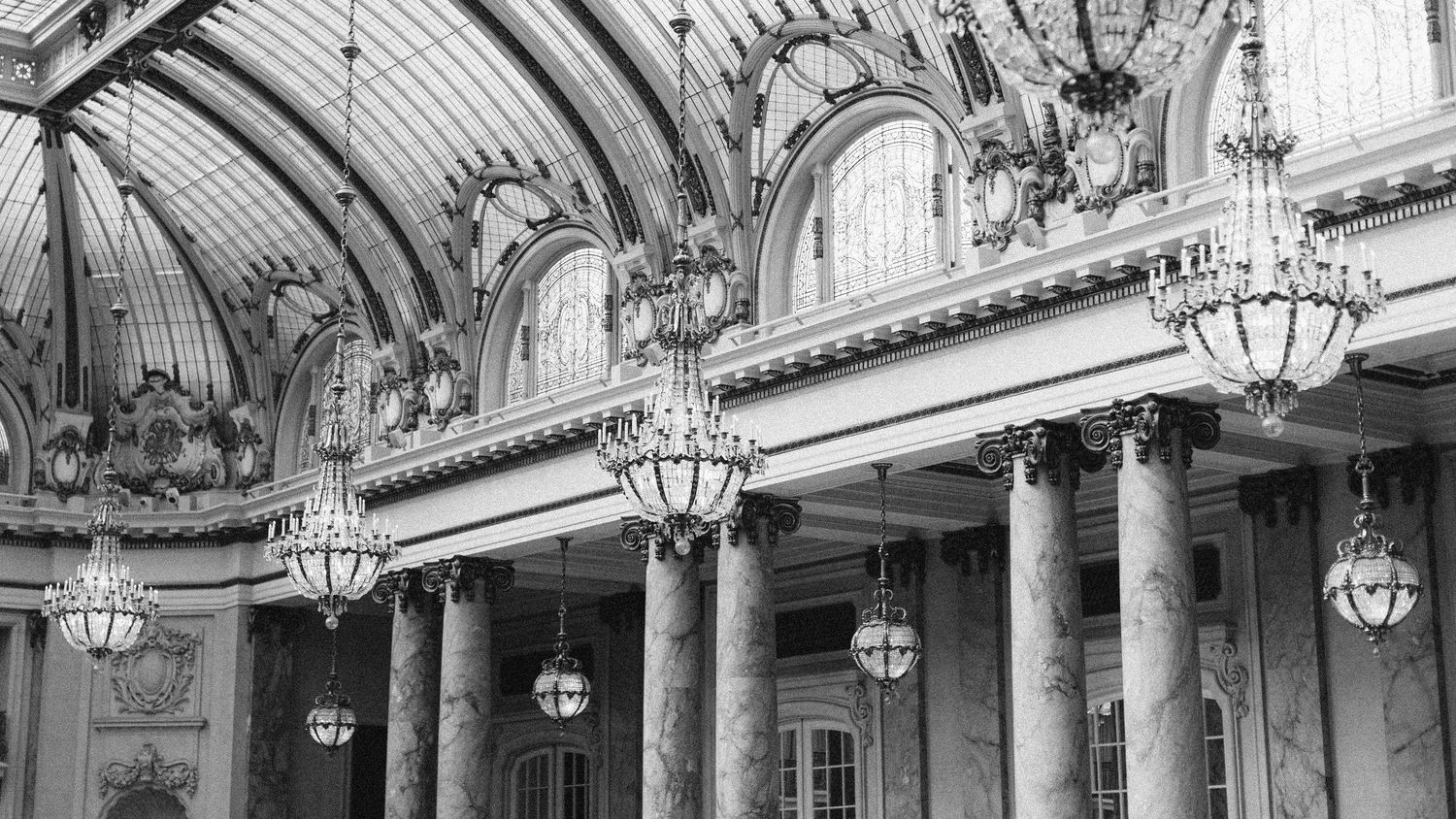 Black and white photo of ornate grand hall with arched glass ceiling, chandeliers, and decorative columns.