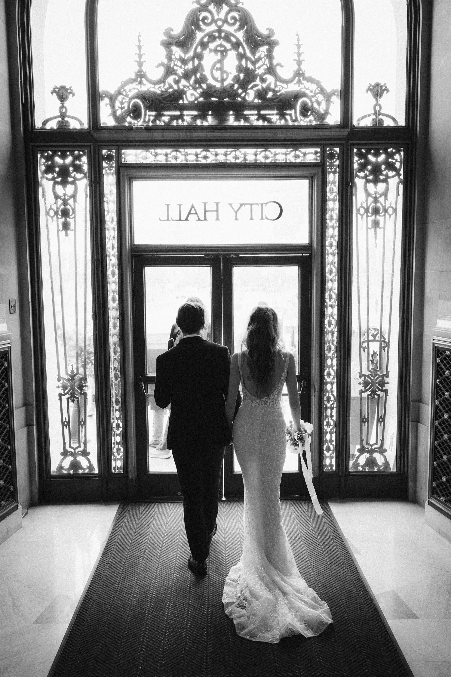 Black and white photograph of a couple in formal wear walking towards elegant glass doors with ornate ironwork designs.